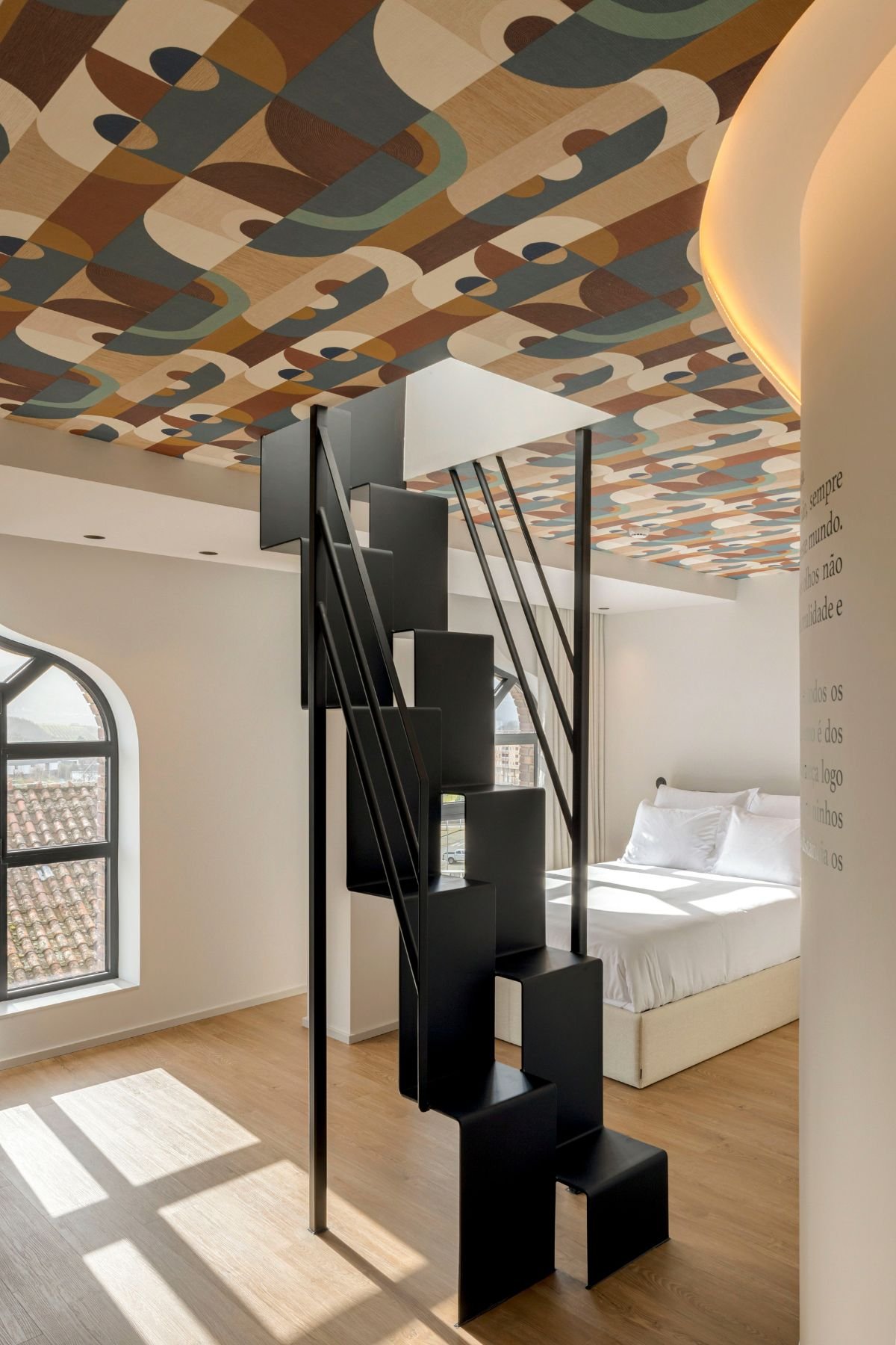 Perspetiva ampla focada no design arrojado da escada assimétrica preta em direção ao piso superior, com enquadramento da cama ao fundo e o padrão do teto em grande plano.