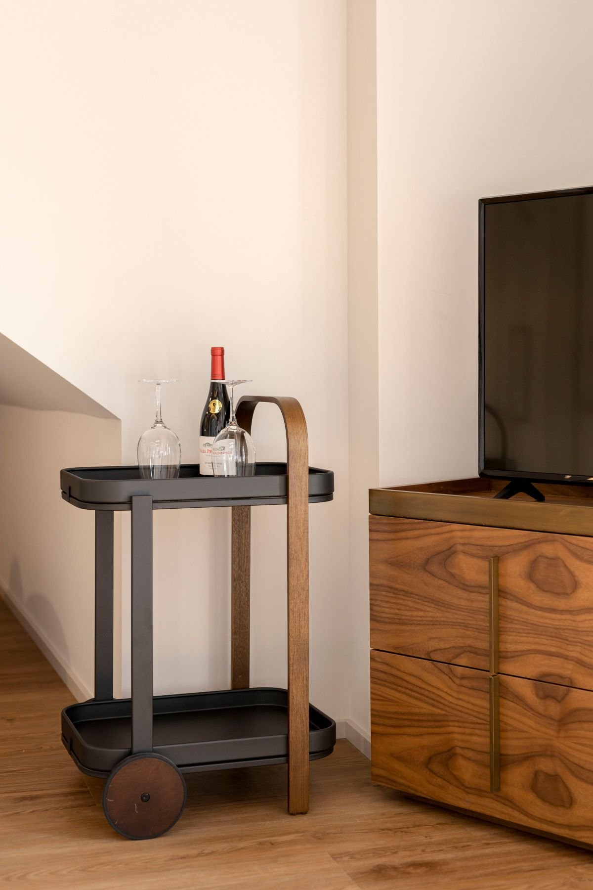 Detalhe do carrinho de apoio de bar em metal preto e madeira, equipado com garrafa de vinho e copos, posicionado ao lado do móvel em madeira com televisão.