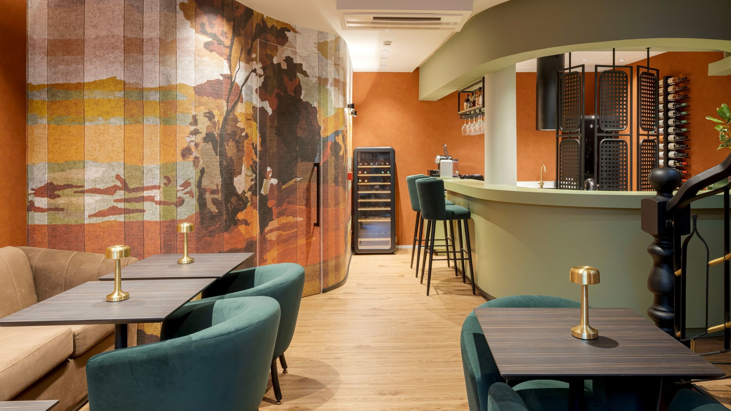 Vista geral do espaço do bar e zona de lounge, integrando mesas com candeeiros dourados, cadeirões em veludo verde escuro, balcão curvo verde-claro e um mural artístico na parede.