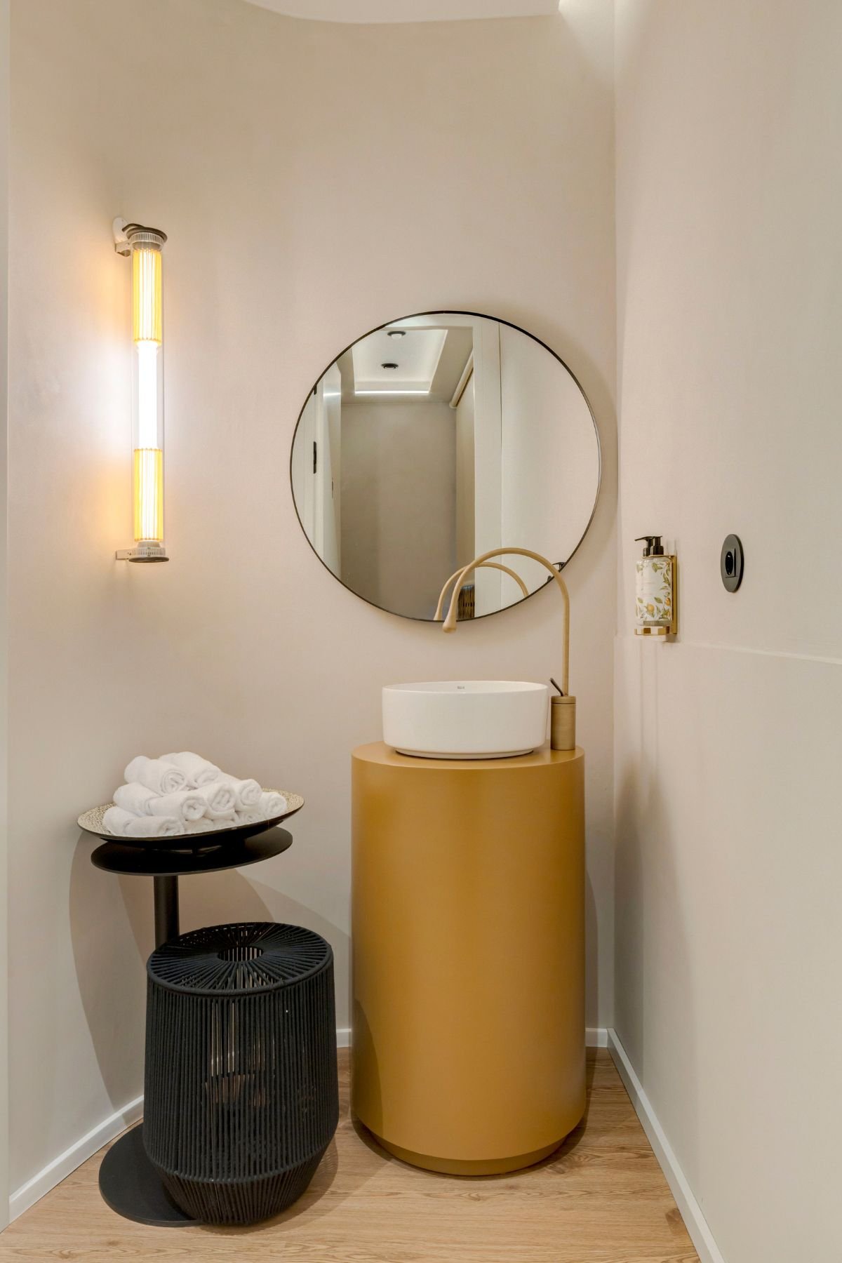 Instalação sanitária com lavatório de pousar num elegante móvel cilíndrico amarelo, espelho redondo de rebordo preto e iluminação de parede tubular de tom quente.