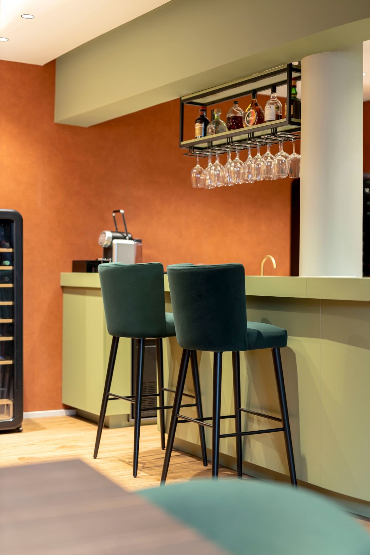 Plano aproximado dos bancos de bar altos em verde escuro junto ao balcão, destacando o suporte suspenso com copos de vinho em vidro.