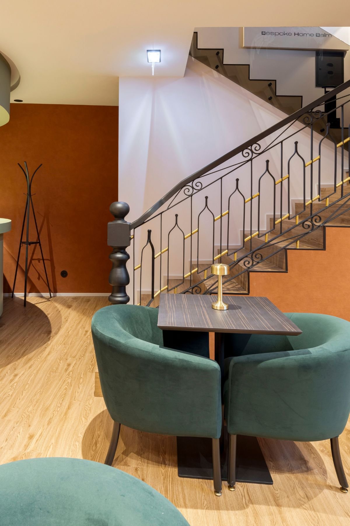 Zona de assento acolhedora com mesa quadrada, cadeiras de veludo verde e candeeiro de mesa dourado, junto à escadaria com guarda-corpo trabalhado em ferro e detalhes dourados.