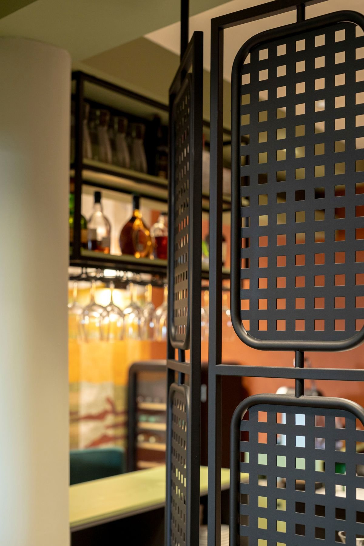 Detalhe em foco da divisória metálica perfurada preta que delimita a área da receção e do bar, com os copos e garrafas visíveis em segundo plano.