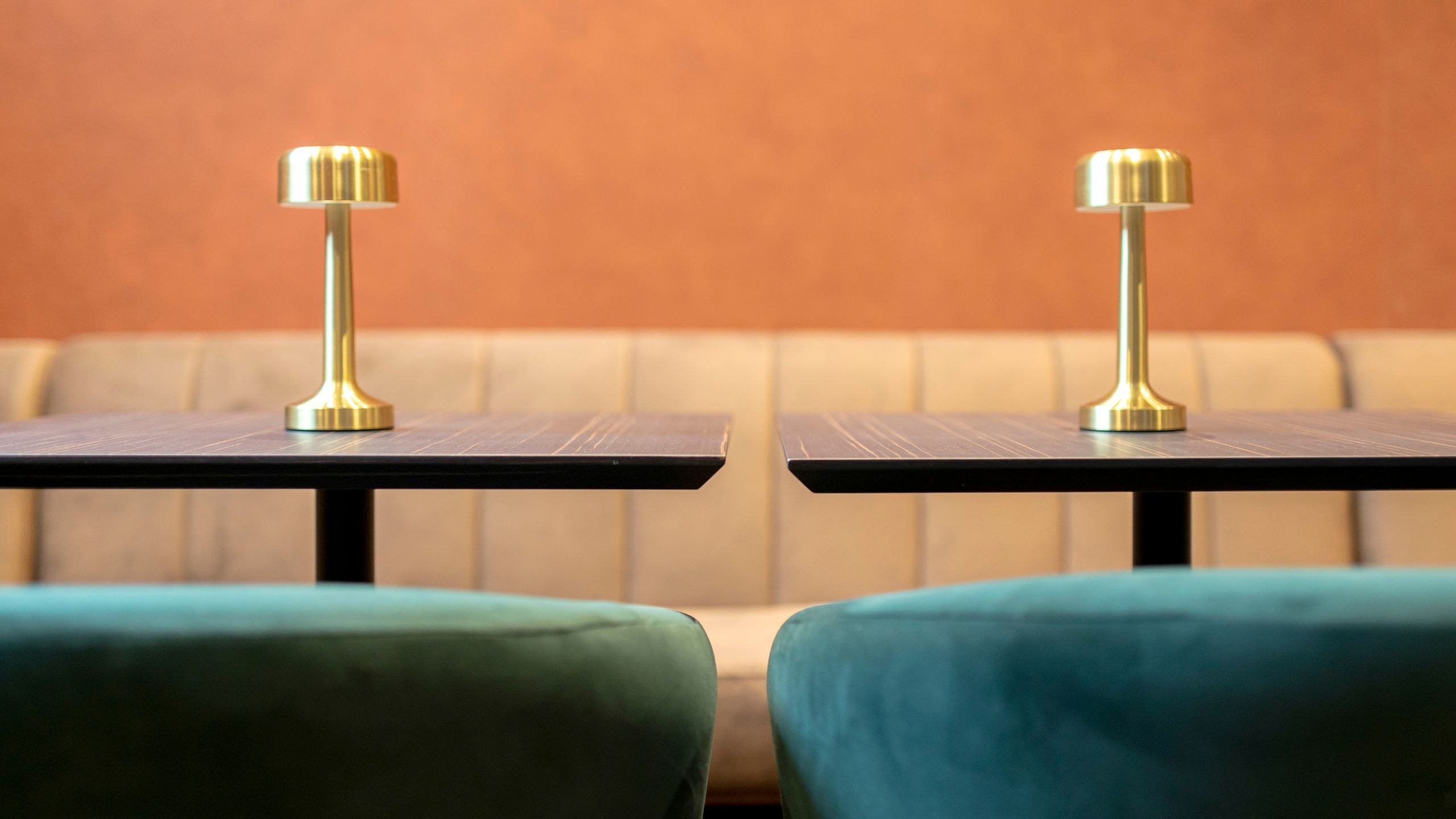 Plano detalhado das mesas escuras com candeeiros de mesa em latão dourado, com o topo dos cadeirões verdes e o sofá estofado ao fundo.