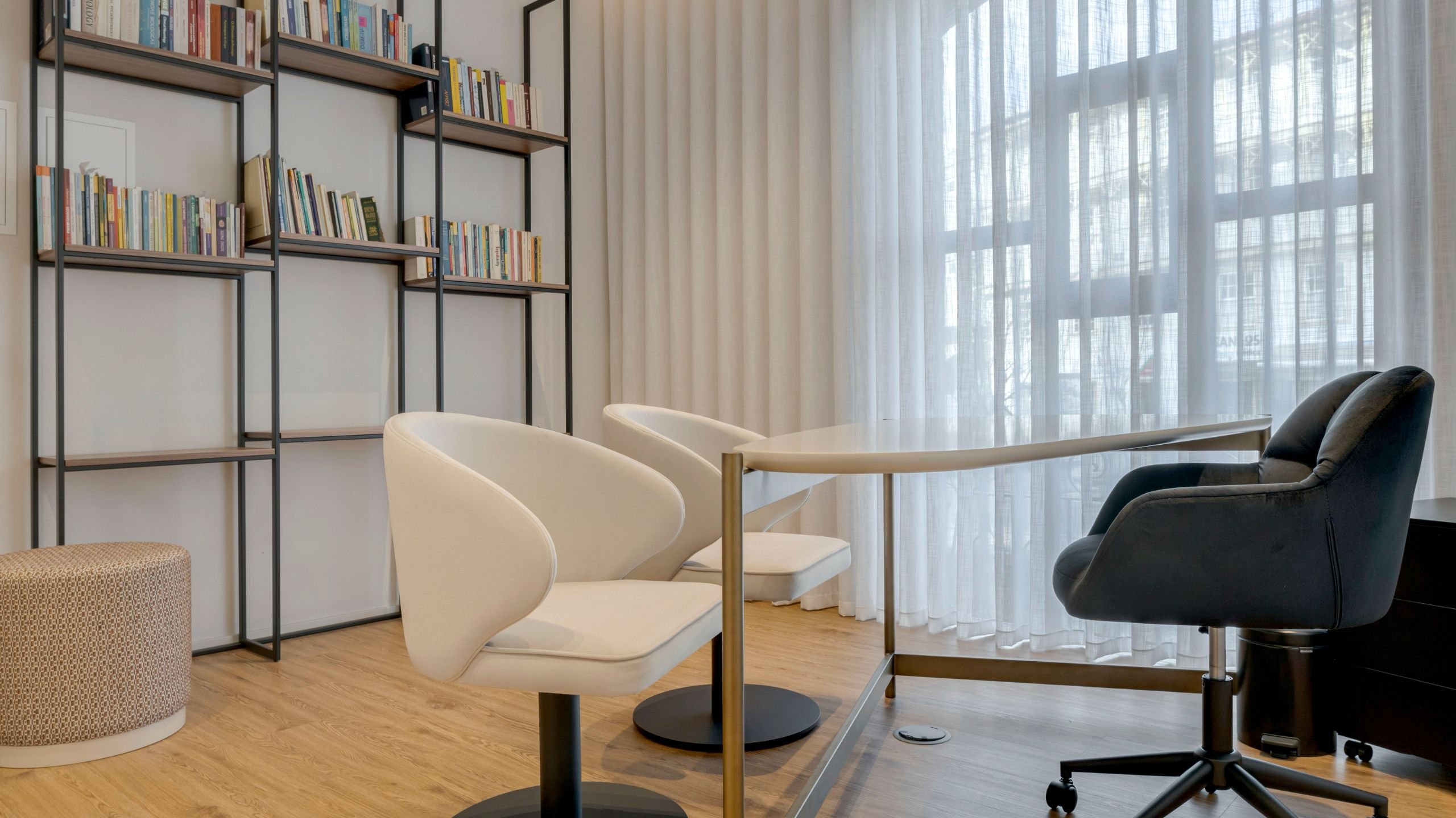 Zona de atendimento do consultório com secretária de tampo claro, cadeiras rotativas em tom marfim para os pacientes, estante metálica preta com livros e puff com padrão discreto.