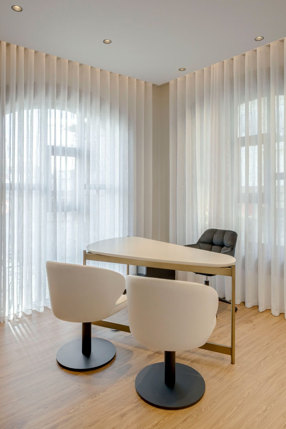 Vista angular da área de consulta iluminada por luz natural através de cortinados fluídos brancos, destacando a secretária de linhas simples, a cadeira do profissional em tom escuro e as duas cadeiras rotativas brancas.