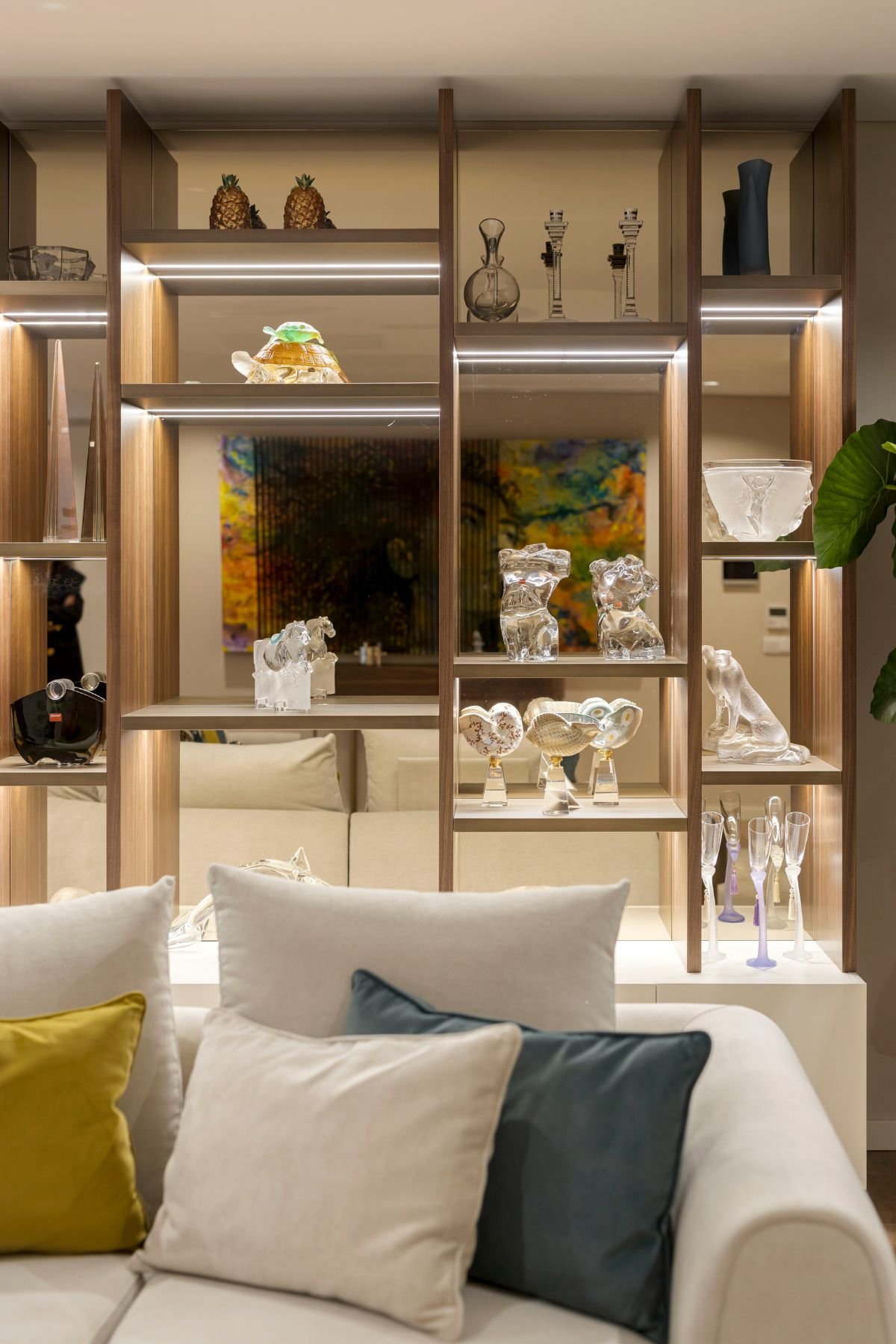 Pormenor de estante em nogueira com nichos iluminados e estatuetas decorativas atrás do sofá claro da sala.