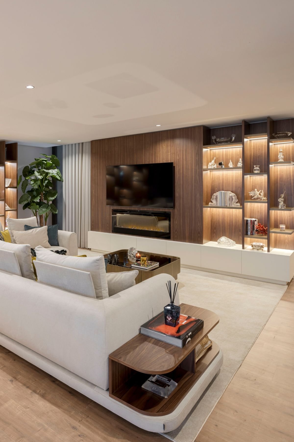 Sala de estar com sofá claro, apoio lateral em nogueira, mesa de centro e painel de TV com lareira.