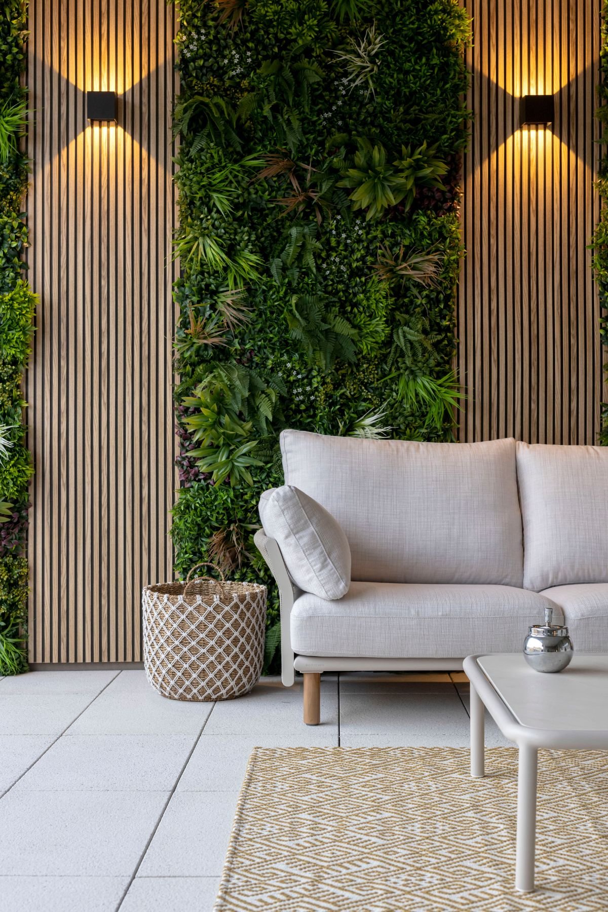 Sofá de exterior claro junto a painel ripado de madeira com vegetação vertical e apliques de luz quentes.
