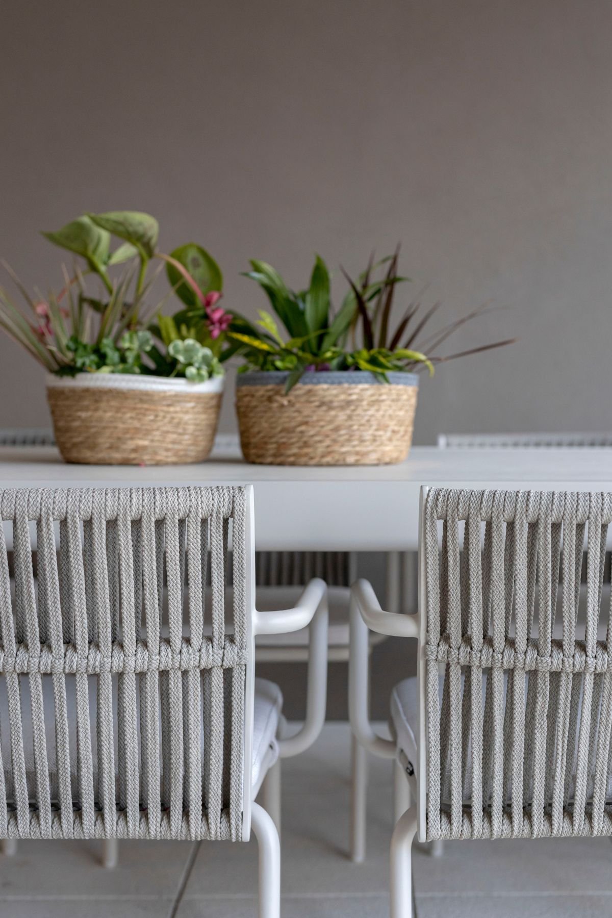 Pormenor das cadeiras de refeição de exterior com trançado cinza e cesto decorativo com plantas sobre a mesa.