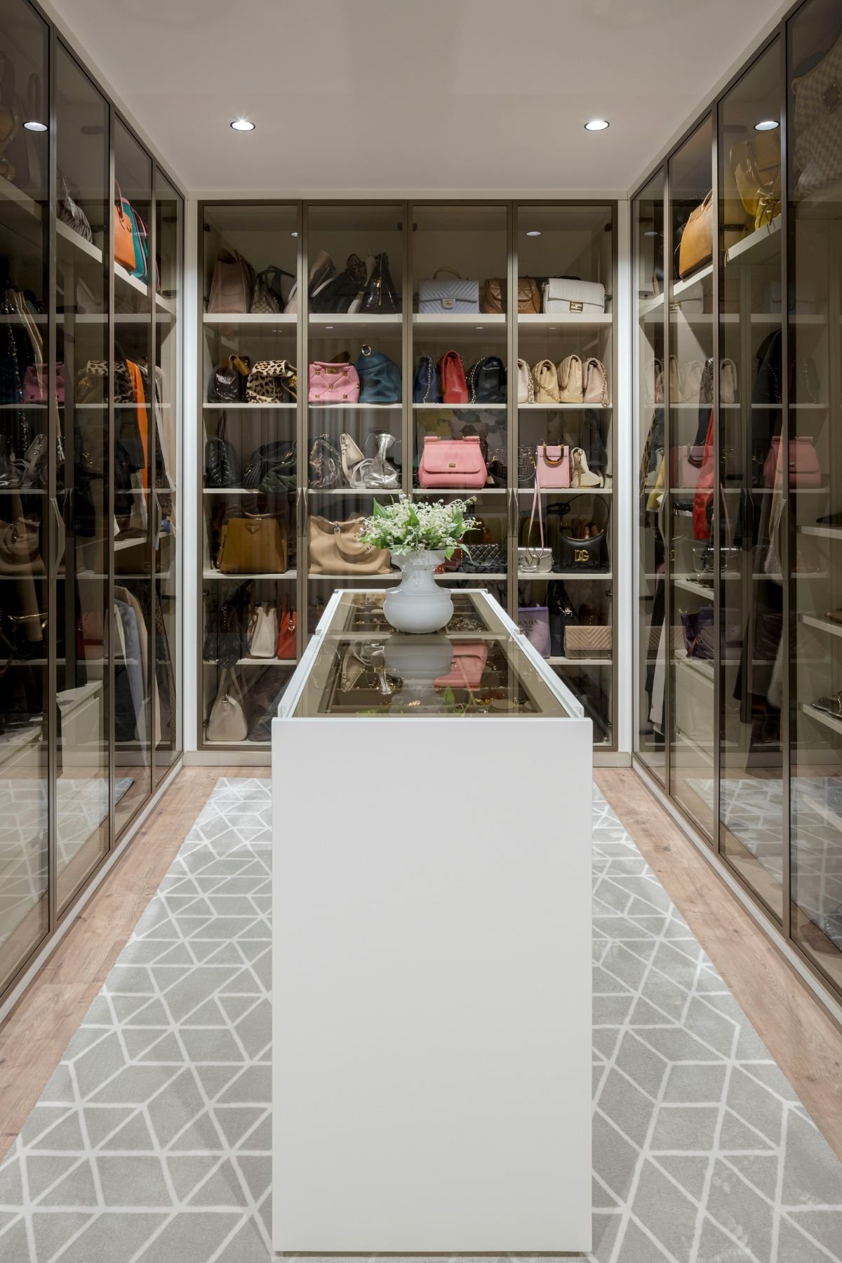 Closet luxuoso com armários iluminados de portas de vidro, ilha central em branco mate com tampo em vidro e tapete de padrão geométrico.