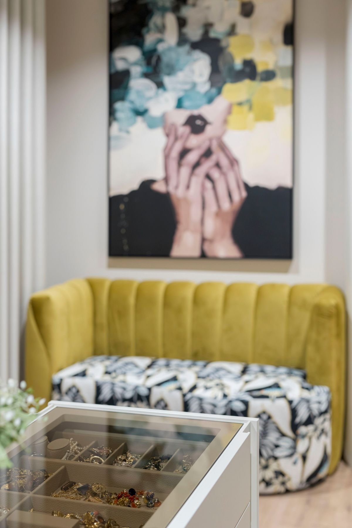 Pormenor do tampo de vidro da ilha de closet com joias expostas, em primeiro plano de um sofá amarelo de veludo e quadro decorativo.