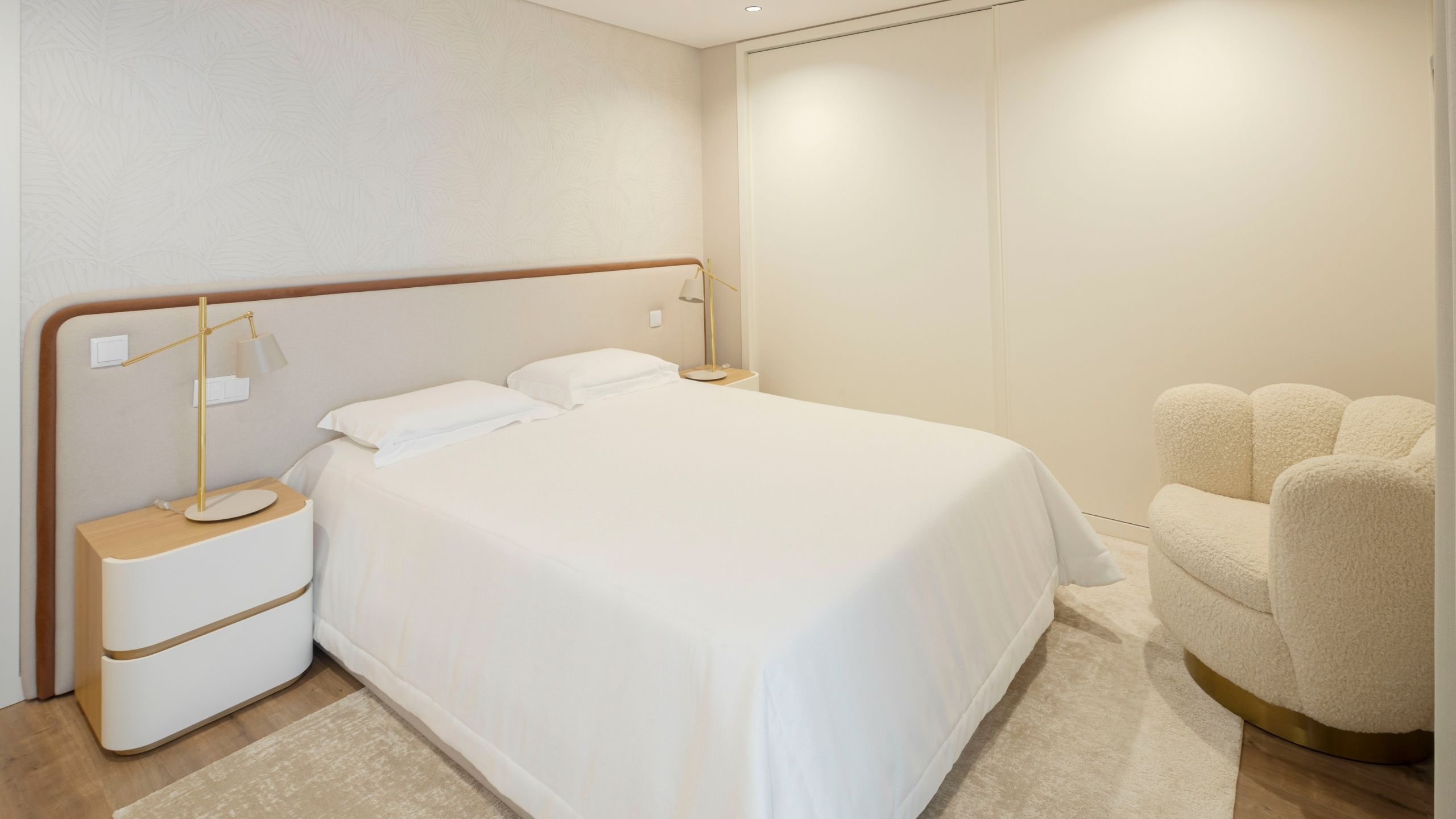 Quarto de hóspedes com cama casal, cabeceira de linhas curvas em tom neutro, mesas de cabeceira com topo em madeira e poltrona estofada bouclé em tom claro.