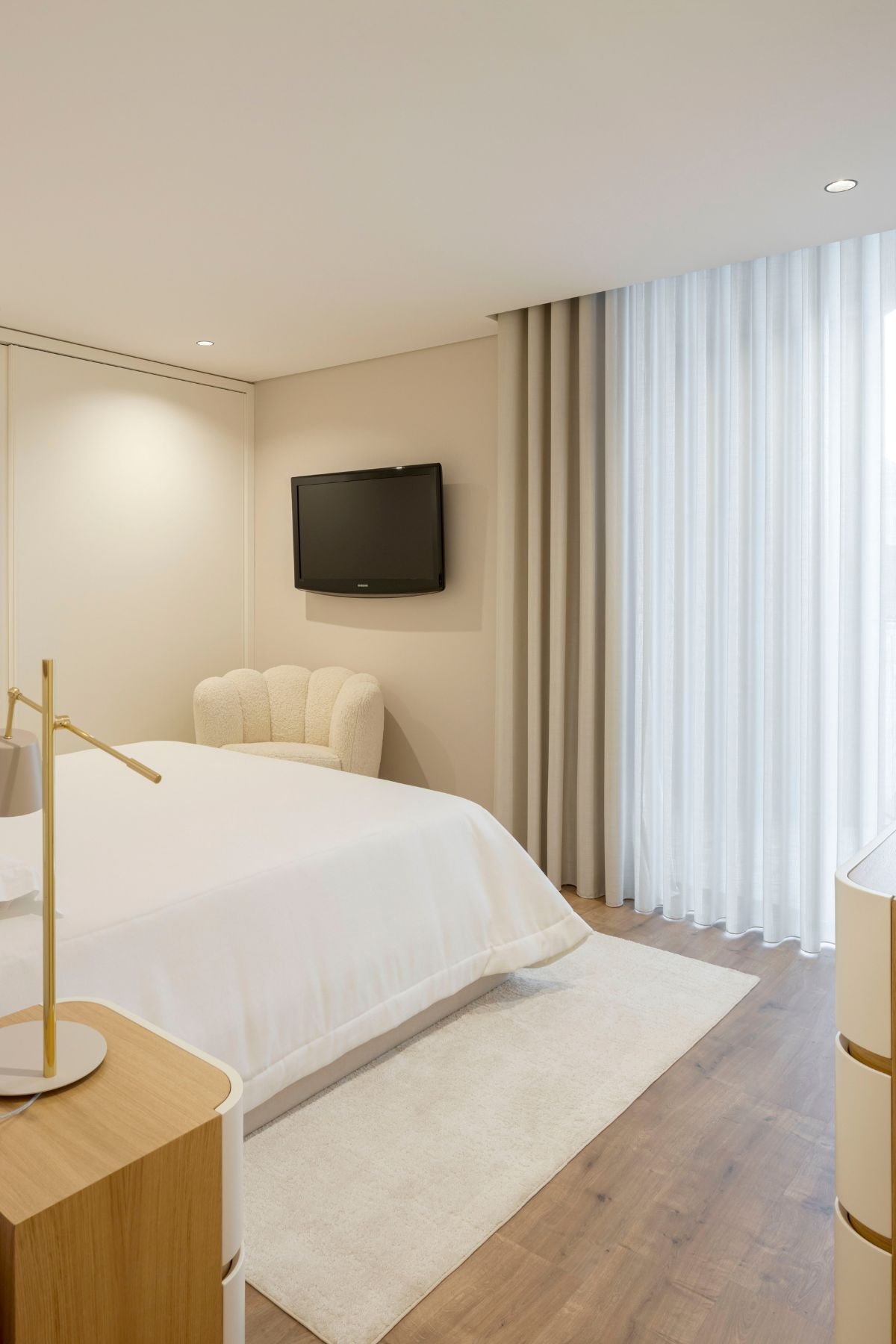 Vista geral do quarto com cama de casal, poltrona bouclé, televisão de parede e janelas com cortinas fluídas.