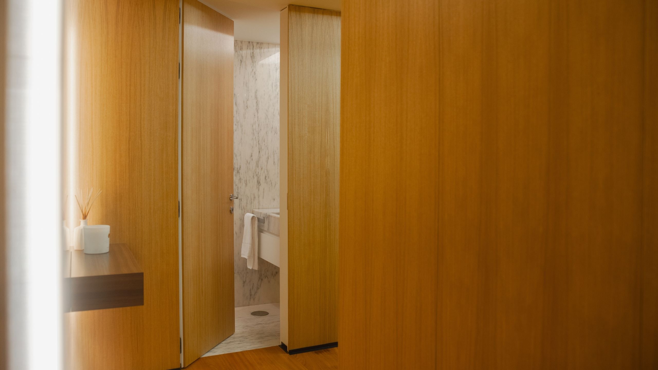 Vista de corredor com painéis de madeira natural e porta parcialmente aberta a dar acesso a uma casa de banho social com lavatório em mármore.