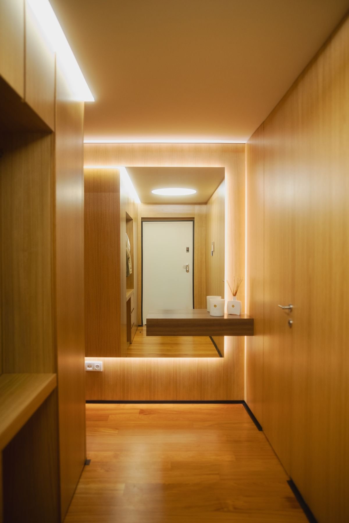 Hall de entrada pequeno revestido a madeira com iluminação quente embutida, espelho de grande dimensão e consola suspensa minimalista.