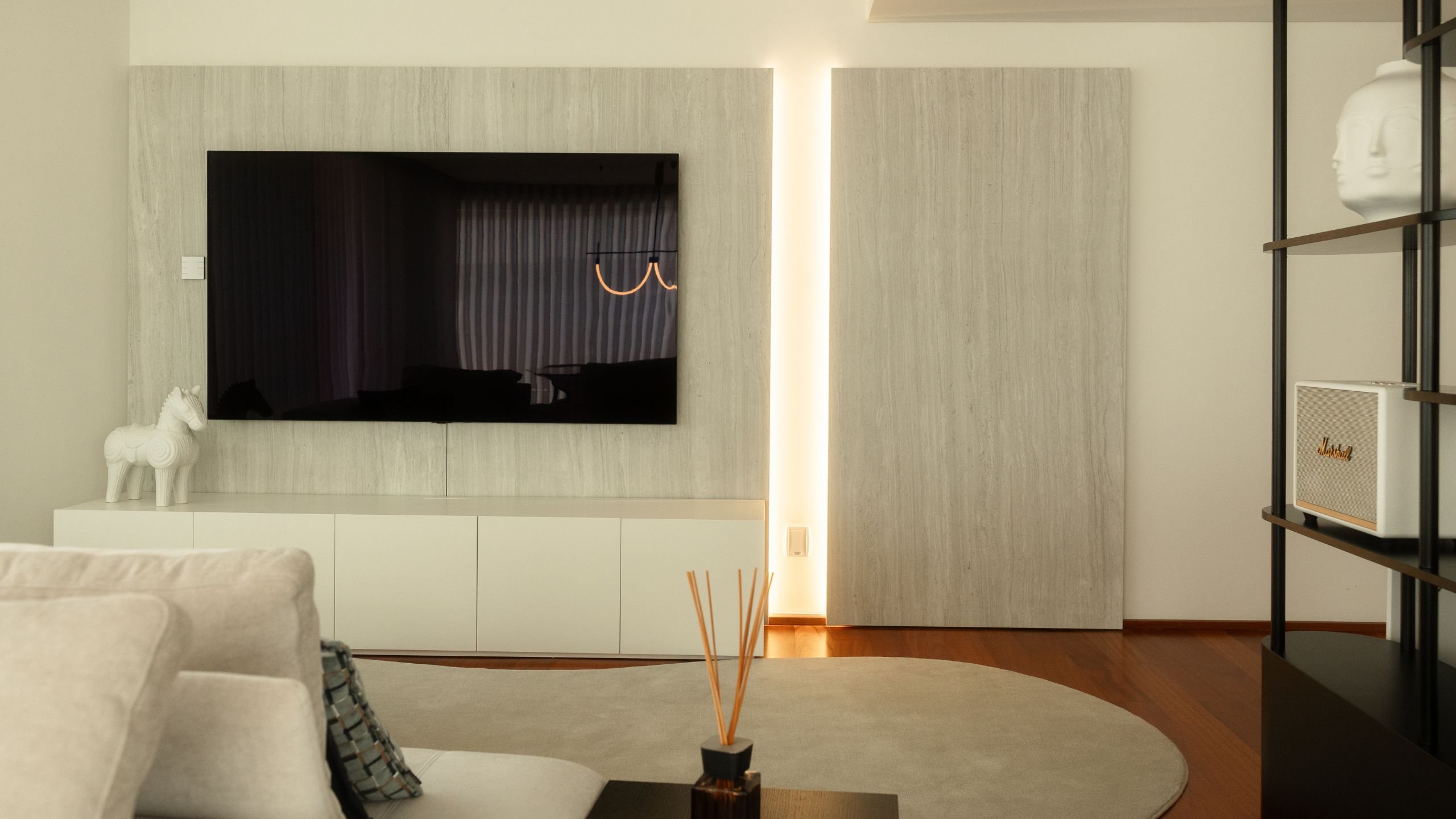 Painel de TV em cerámico com iluminação vertical contínua, móvel inferior branco e estante metálica decorativa.