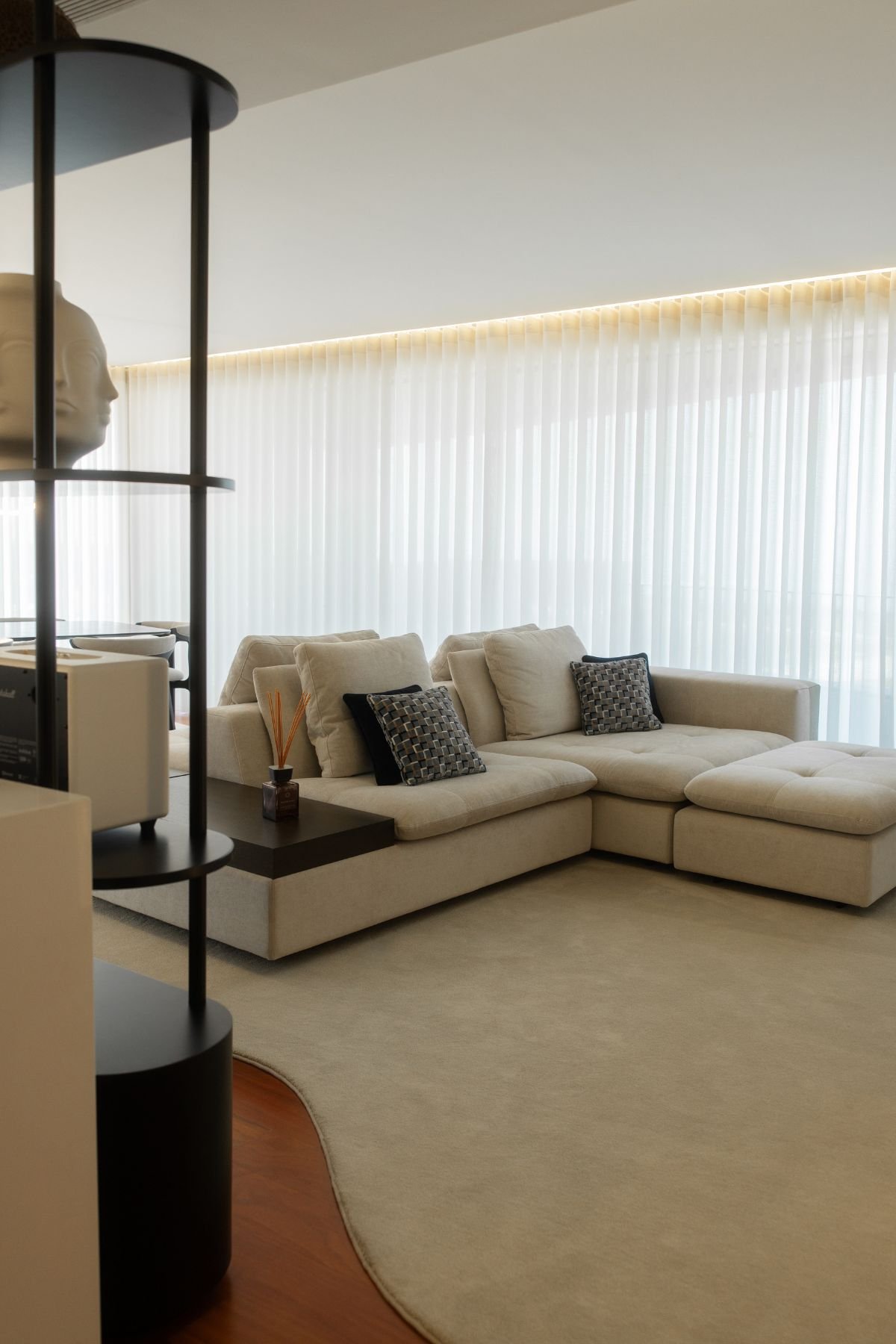 Perspetiva da sala de estar em open space com sofá modular bege, tapete amplo em tons neutros e cortinas brancas ao fundo.
