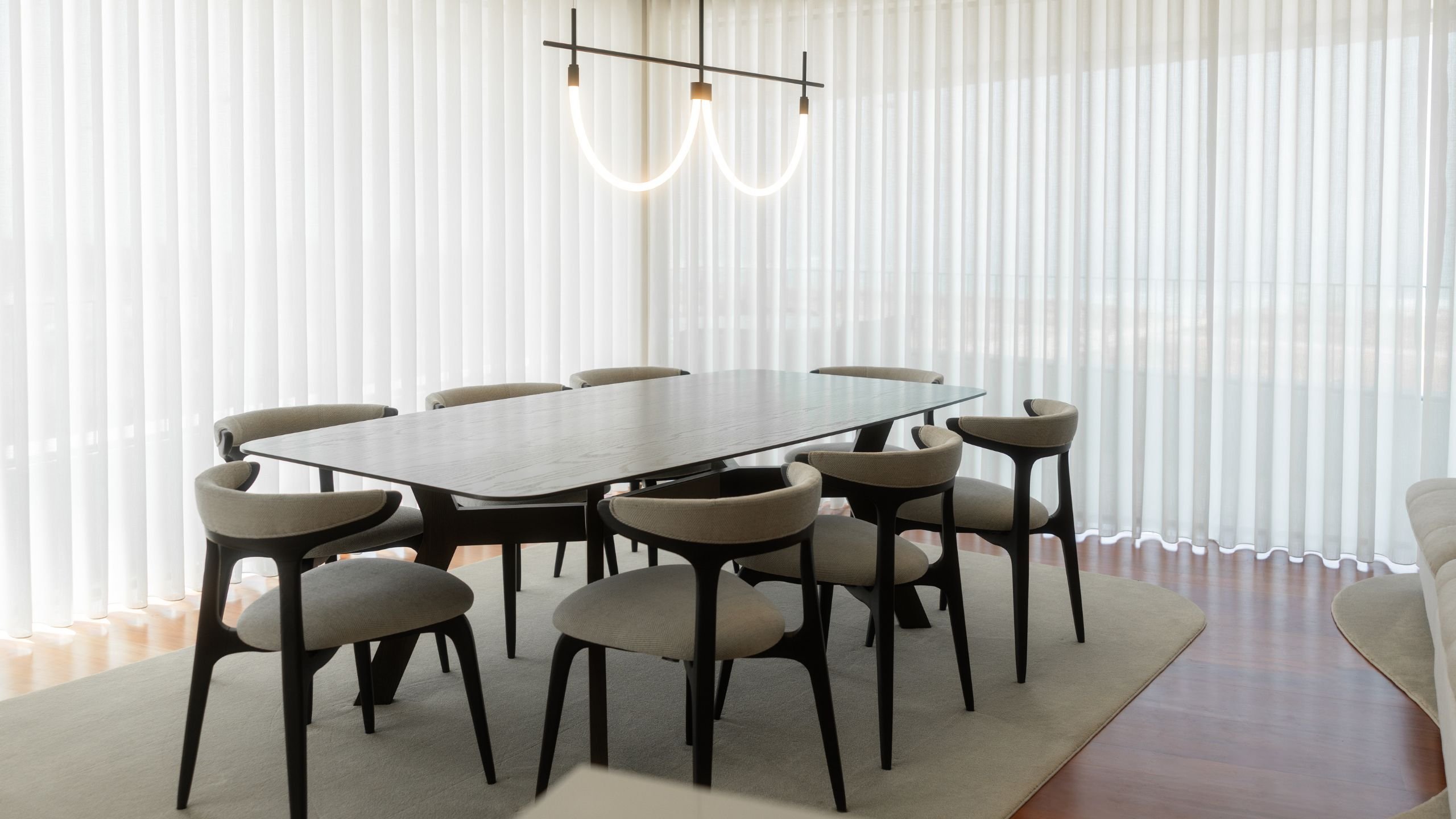Sala de jantar moderna com mesa retangular em madeira escura, cadeiras estofadas e candeeiro suspenso de design tubular curvo.