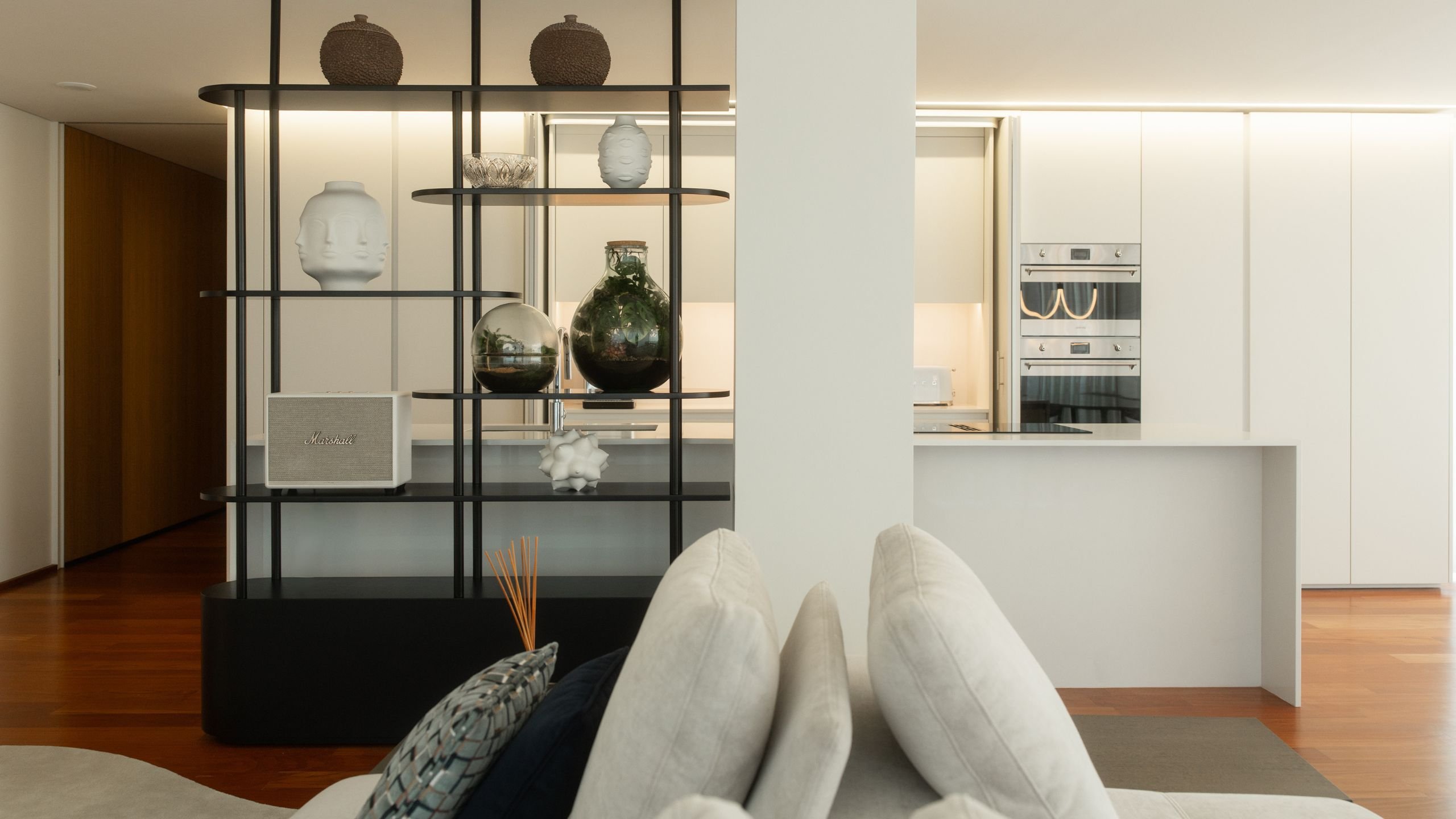 Transição entre a sala de estar e a cozinha moderna através de estante metálica preta vazada com objetos decorativos.