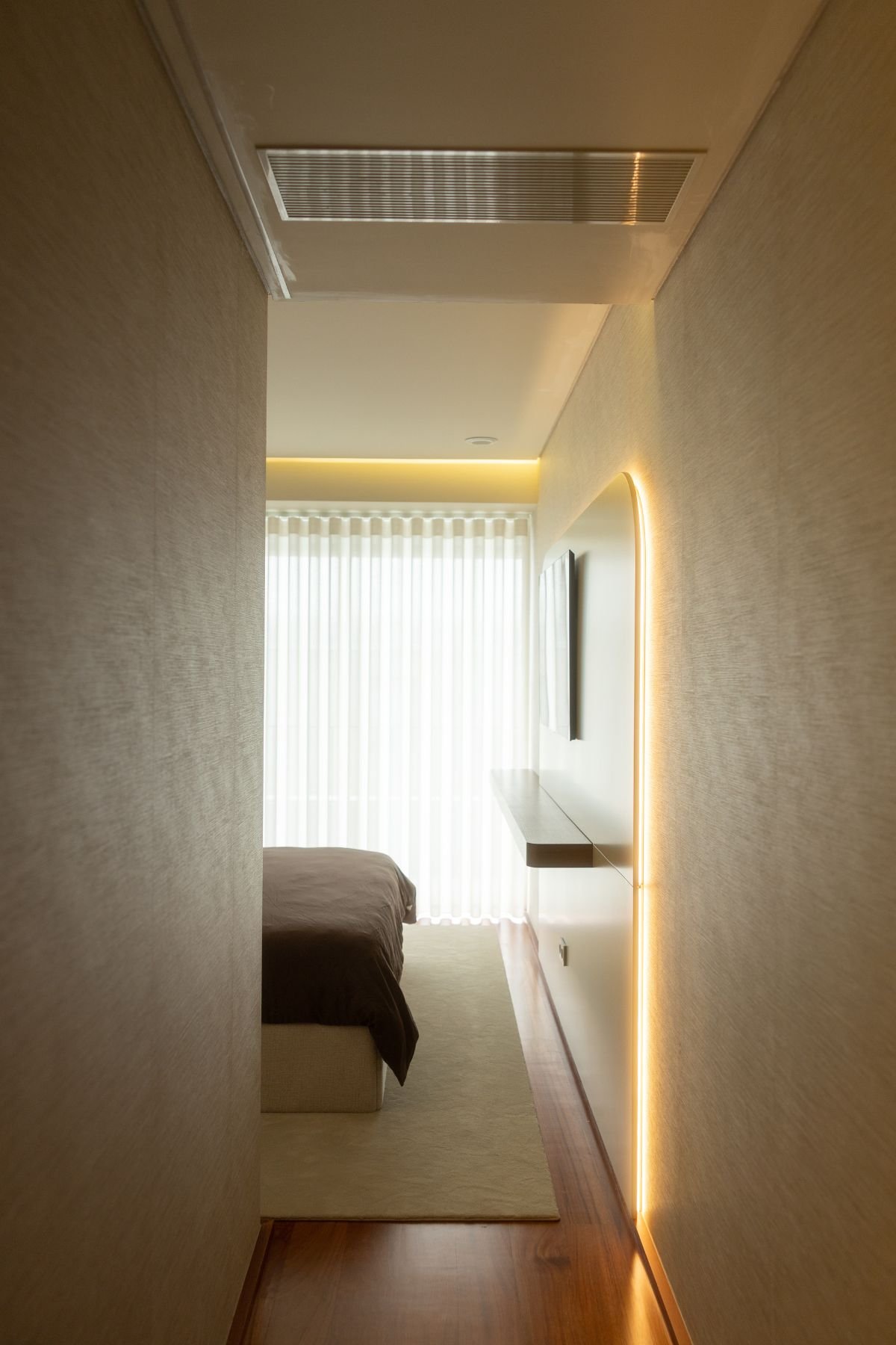 Perspetiva de corredor de acesso ao quarto com painel de TV iluminado por luz LED indireta.