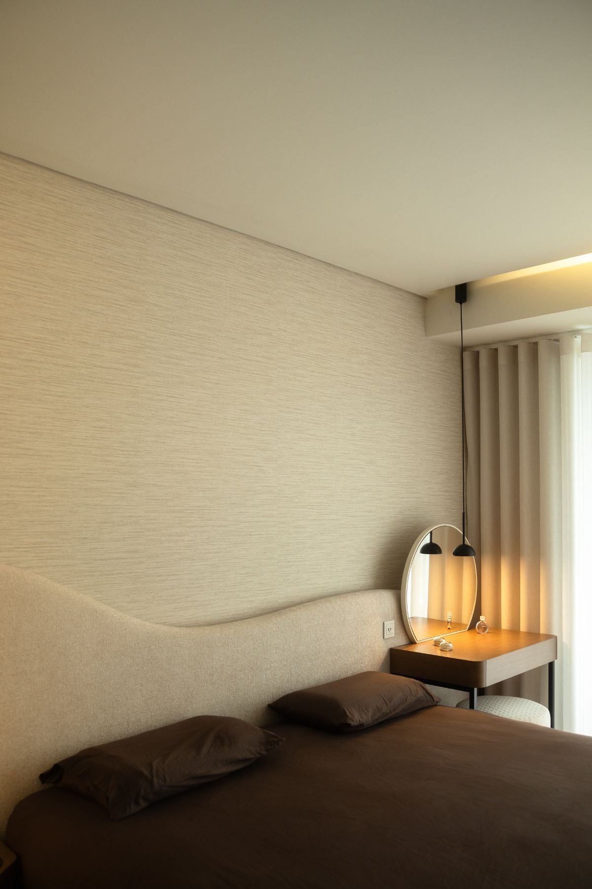 Vista superior da cama de casal com cabeceira de linhas orgânicas e toucador minimalista com espelho redondo.