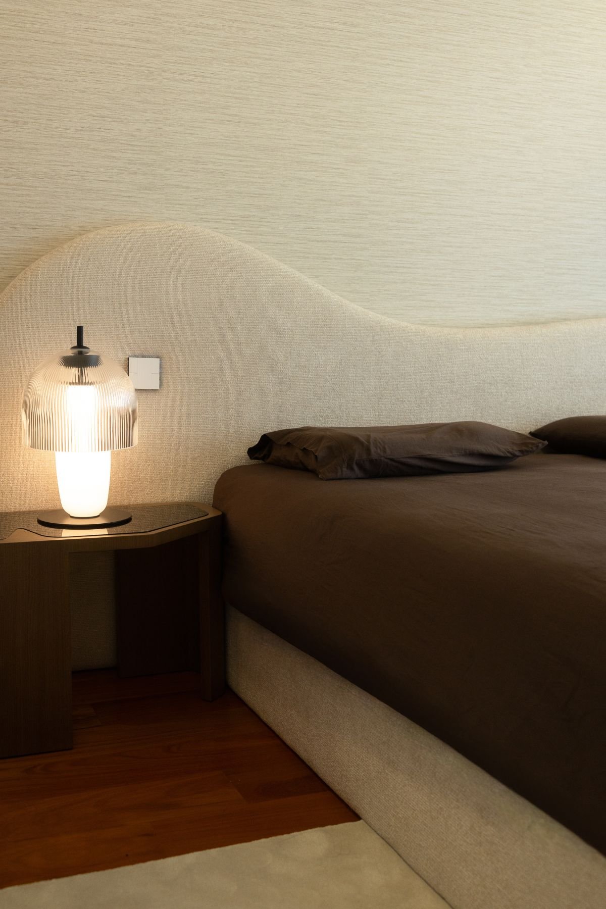 Pormenor da mesa de cabeceira em madeira escura com candeeiro de vidro raiado aceso ao lado da cama de casal.