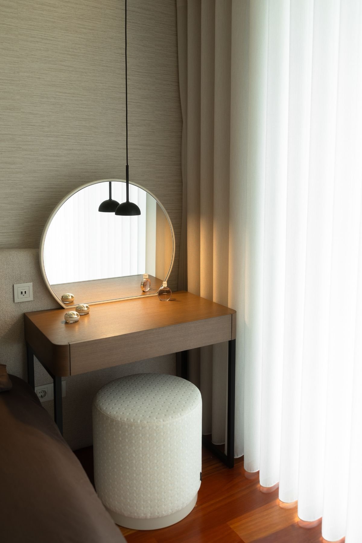 Toucador moderno posicionado junto às cortinas brancas, com espelho redondo iluminado por pendente preto.