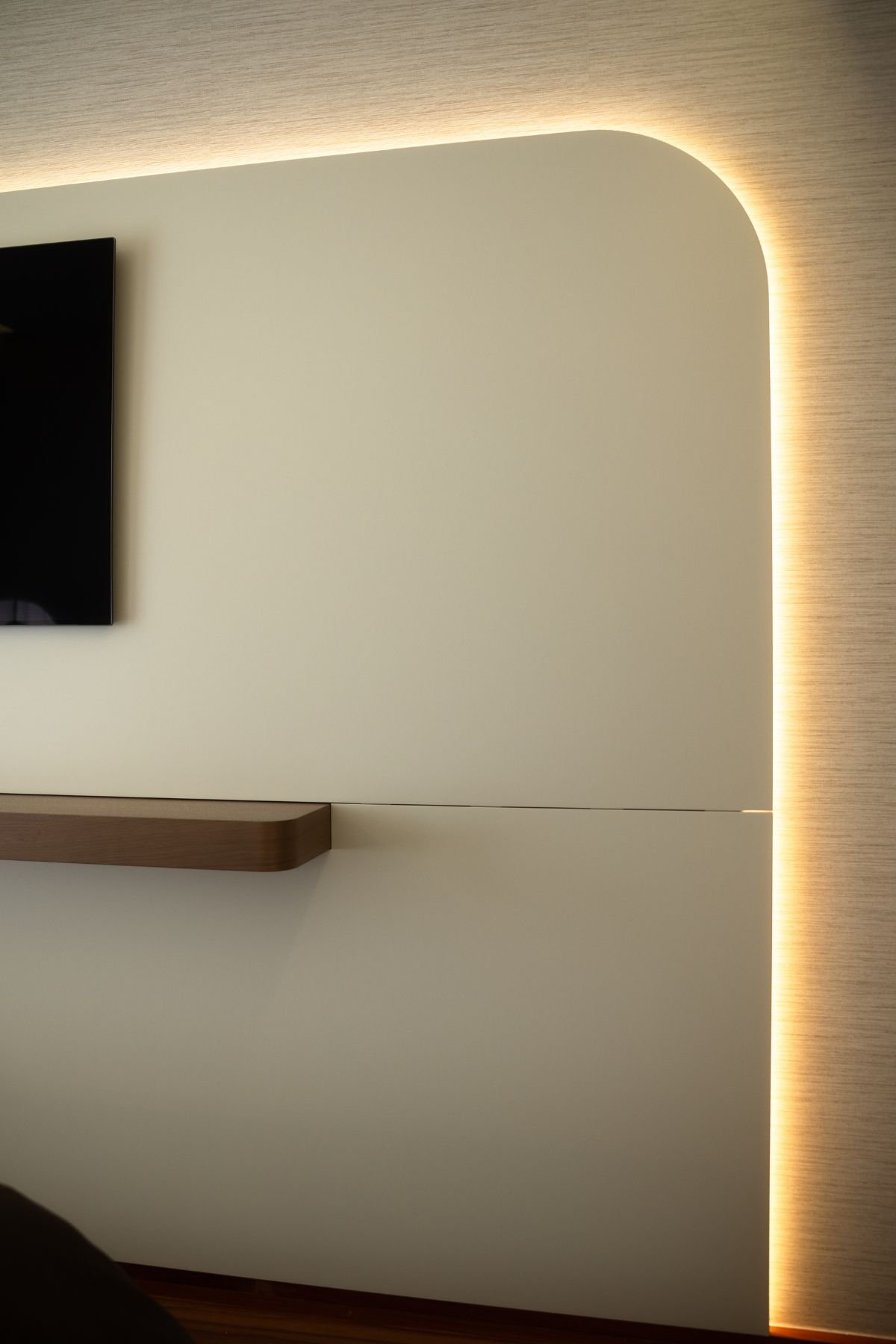 Detalhe do canto arredondado do painel branco de TV com retroiluminação LED contínua e prateleira suspensa em madeira.