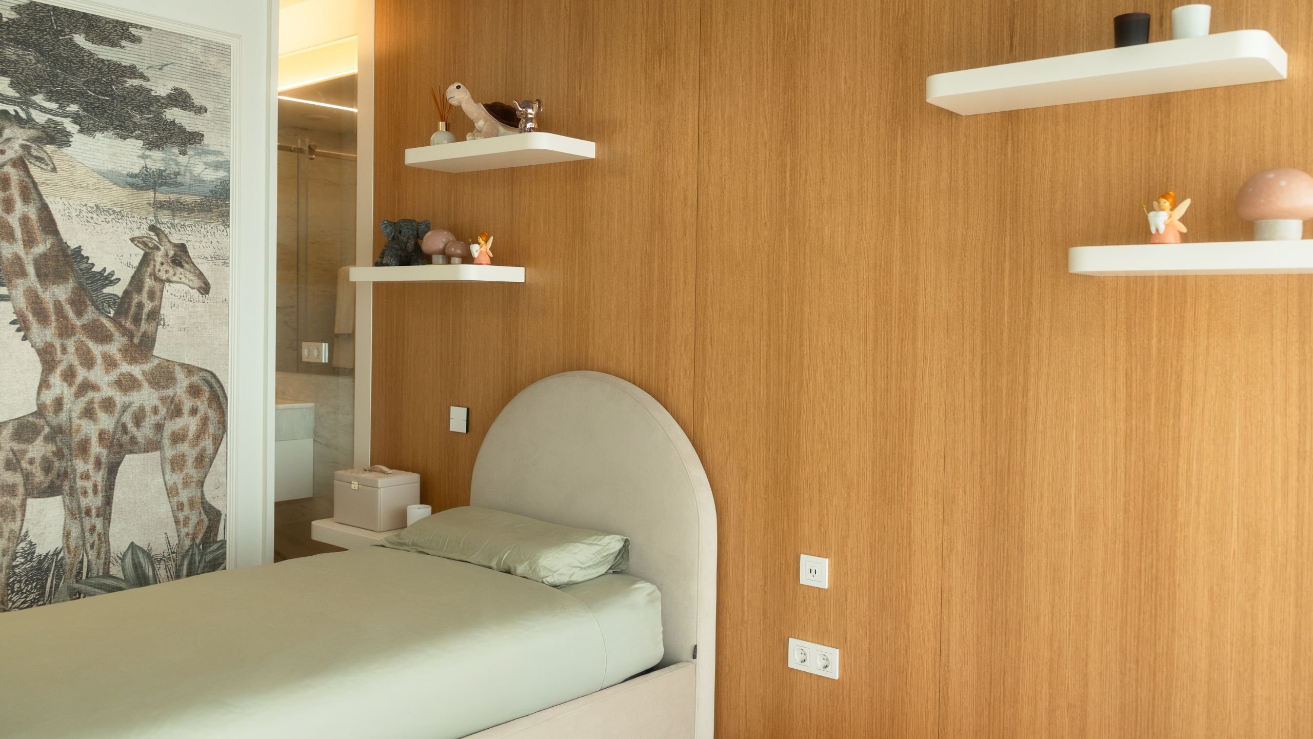 Quarto de criança com painel em madeira, cabeceira estofada arqueada, prateleiras flutuantes com brinquedos e papel de parede com girafas.
