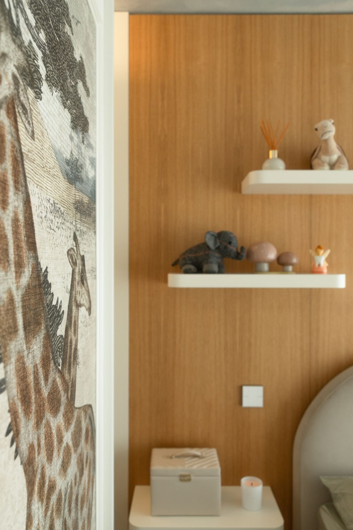 Pormenor do canto do quarto de criança com mesa de cabeceira branca suspensa, prateleiras com peluches e painel de girafas.