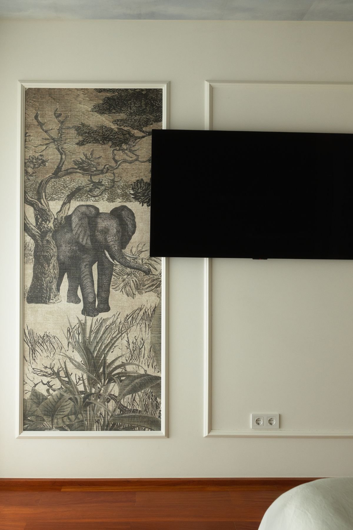 Parede branca com molduras decorativas em boiserie enquadrando painel impresso com elefante e televisão encastrada.