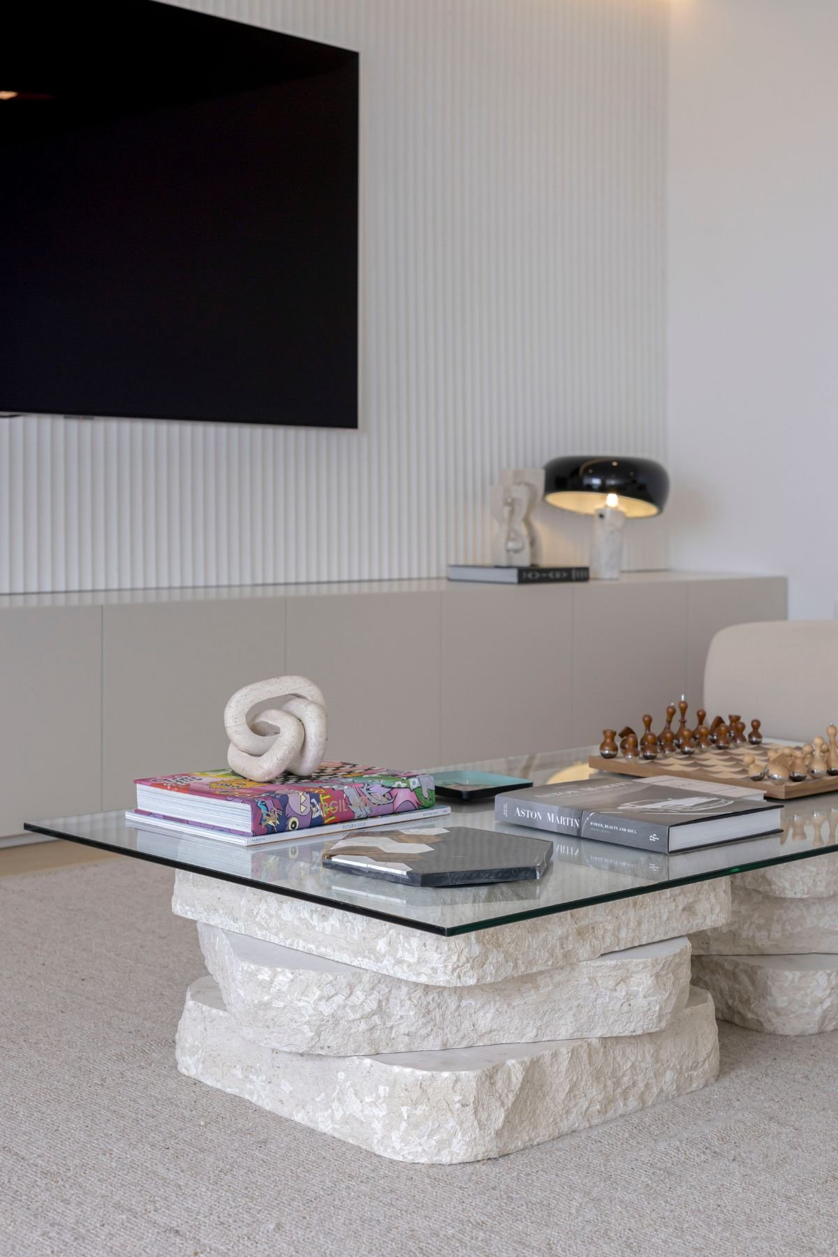 Mesa de centro com tampo de vidro e base em pedra natural esculpida, decorada com jogo de xadrez e livros de design.