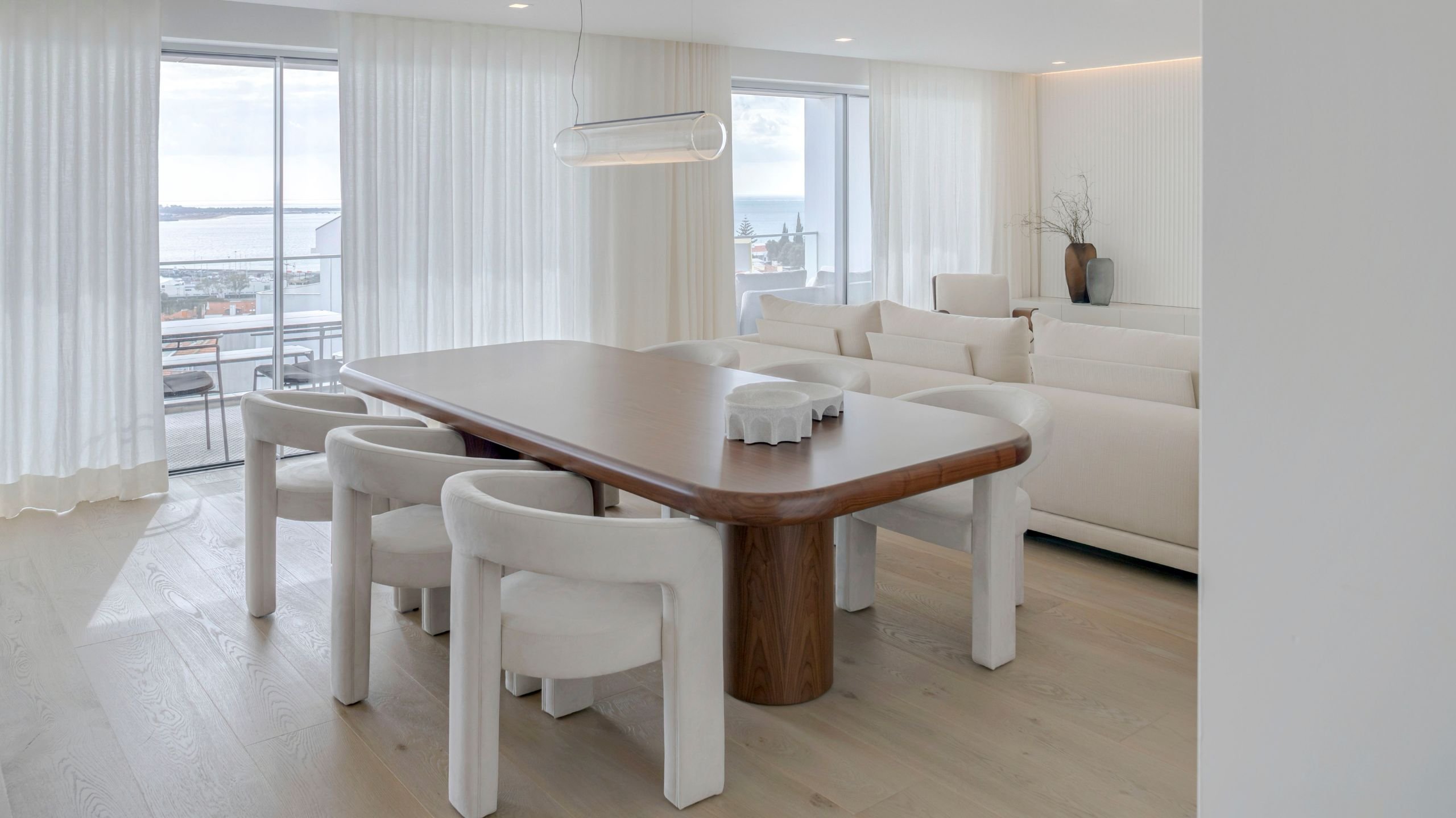 Decoração da sala de jantar em tons neutros, destacando a mesa em madeira, cadeiras estofadas e candeeiro suspenso tubular.