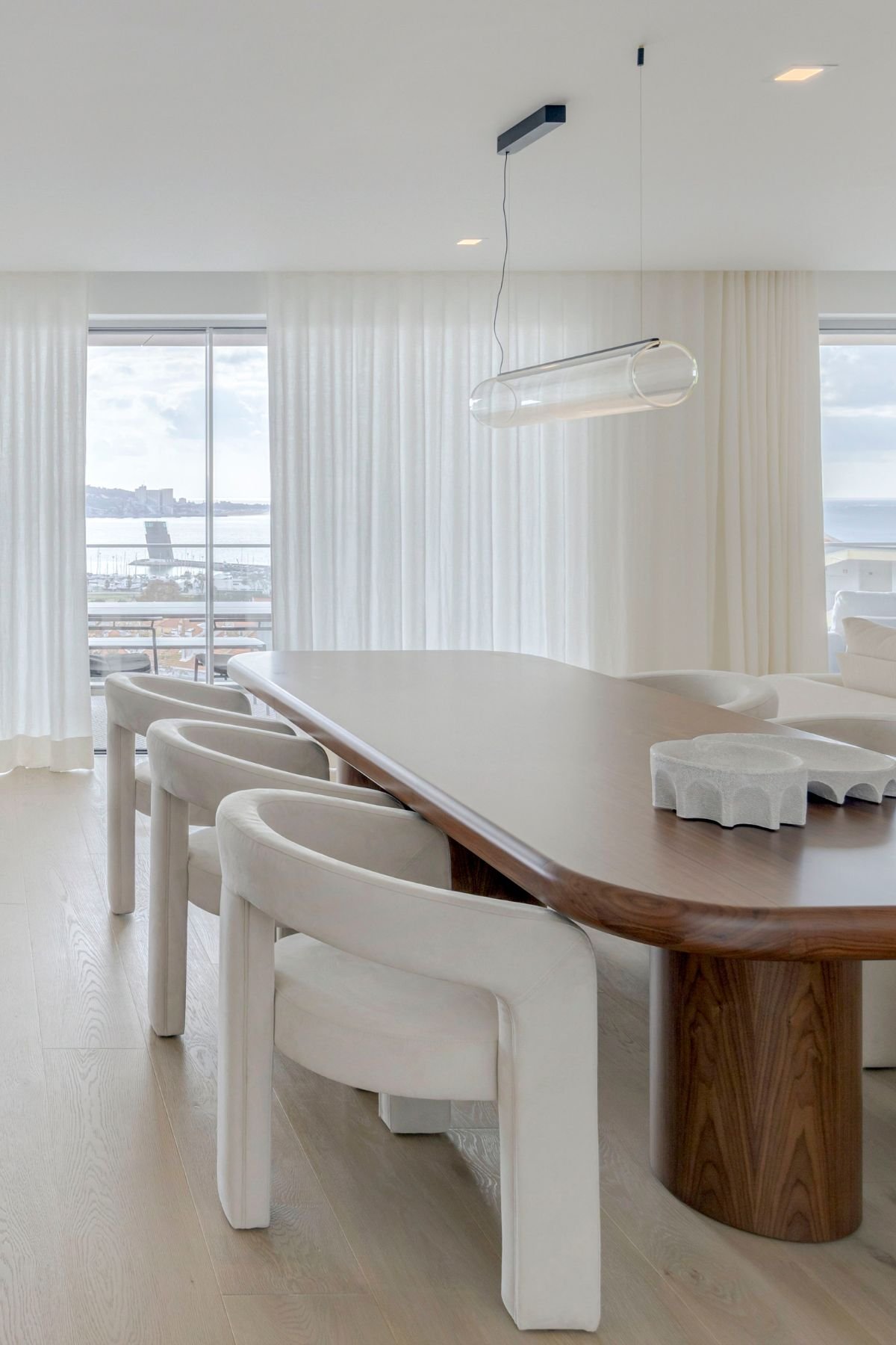 Mesa de jantar em madeira com extremidades arredondadas, rodeada por cadeiras estofadas em tom creme, num espaço com vista exterior.