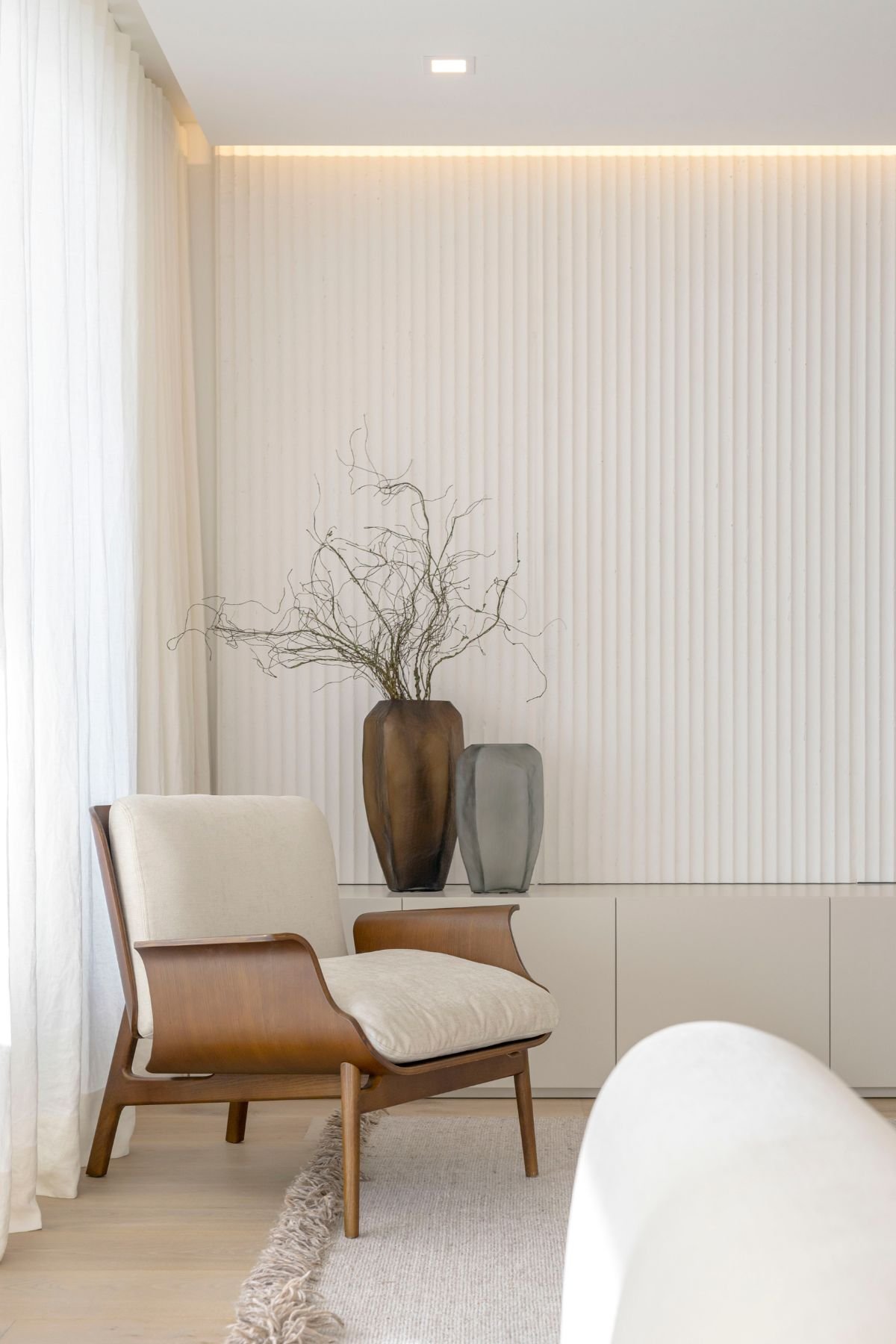 Poltrona de design em madeira e estofos claros junto a jarras decorativas e parede com ripado branco.