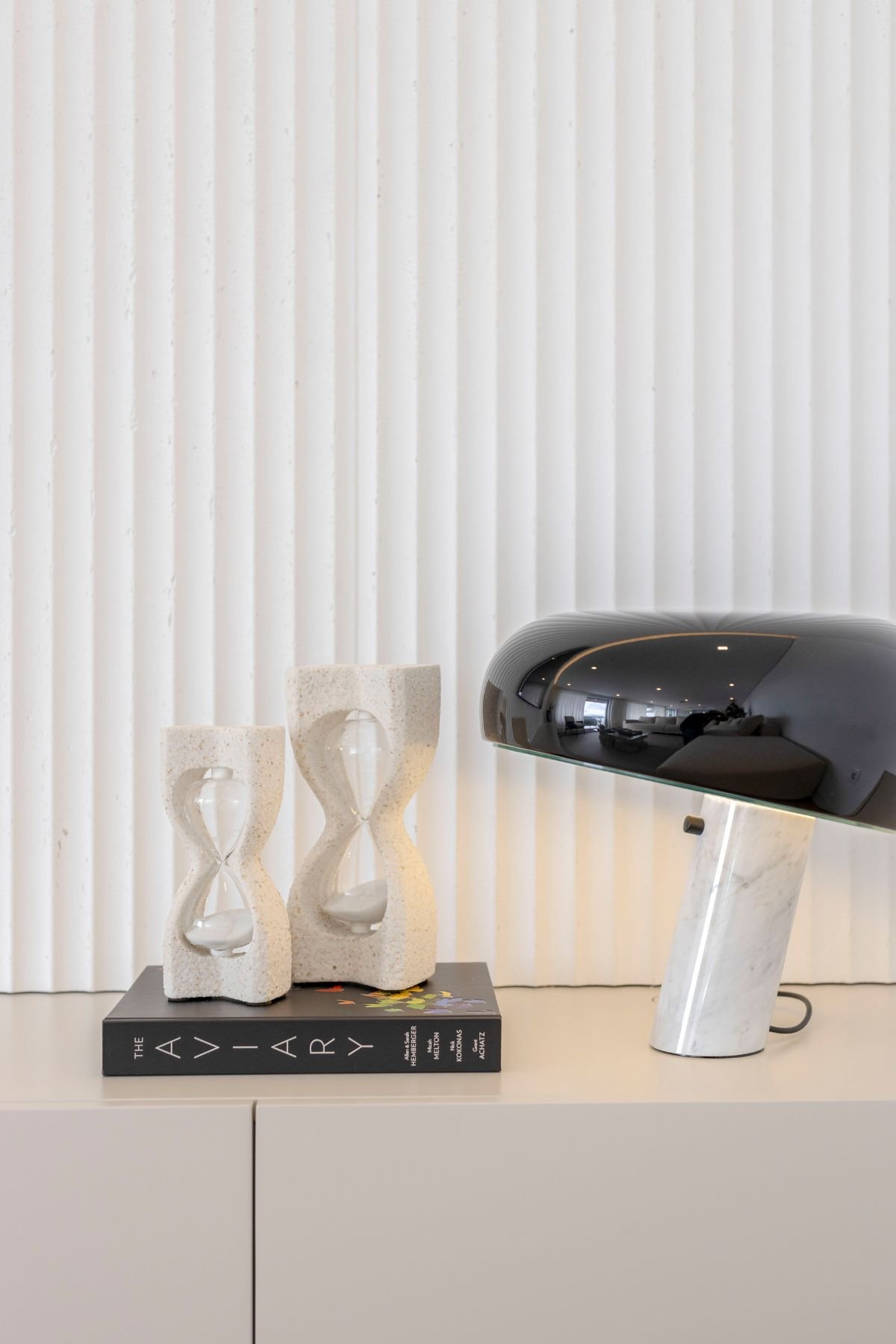 Pormenor decorativo sobre móvel de apoio com ampulhetas em pedra, livro de design e candeeiro de mesa com base em mármore.