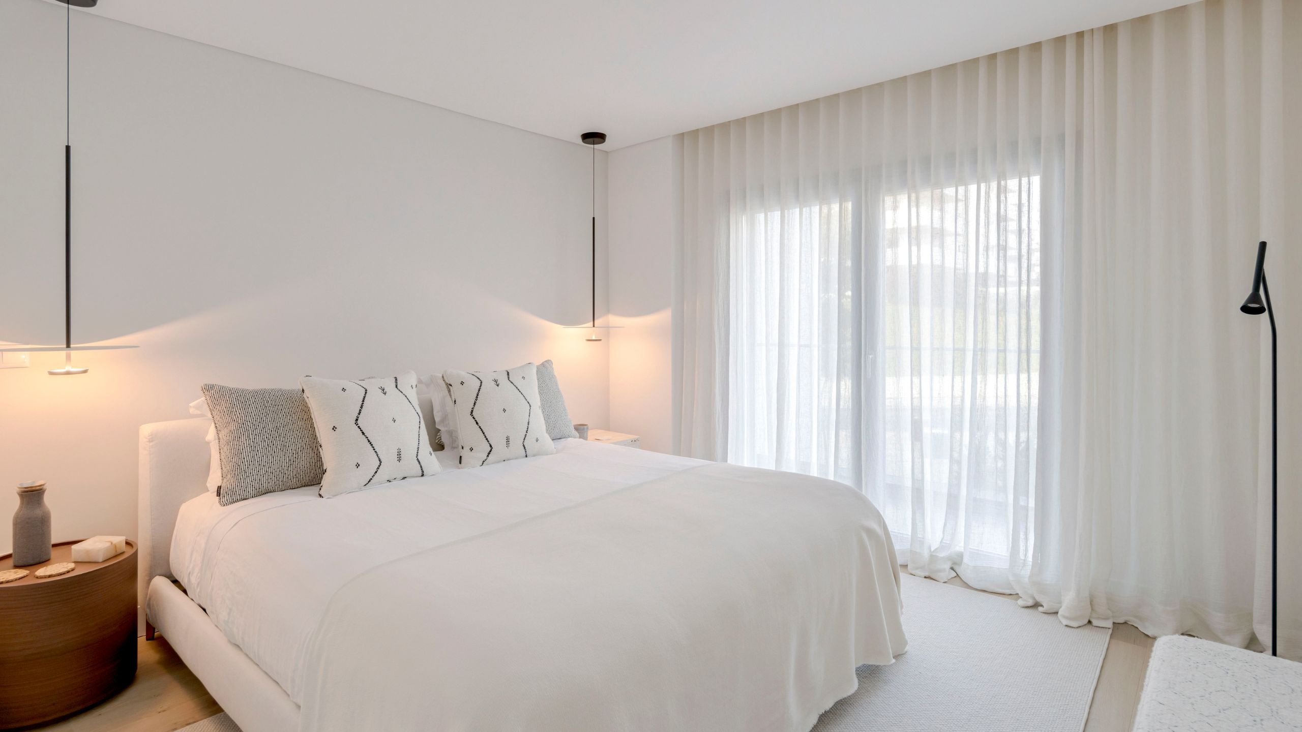 Quarto principal da suite com cama de casal em tons claros, candeeiros suspensos, cortinas de linho e luz natural.