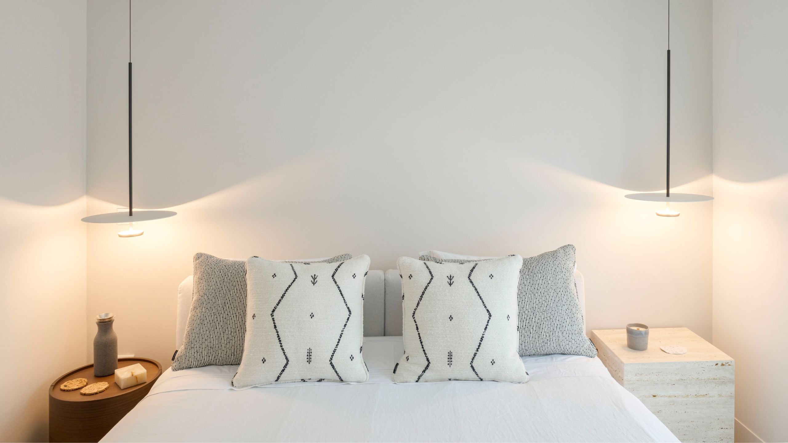 Pormenor da cabeceira de cama com almofadas de padrões étnicos e iluminação suspensa simétrica.