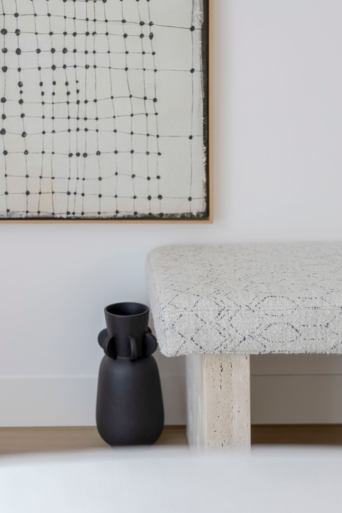 Pormenor de banqueta estofada com pés em pedra Travertino e jarra decorativa preta.
