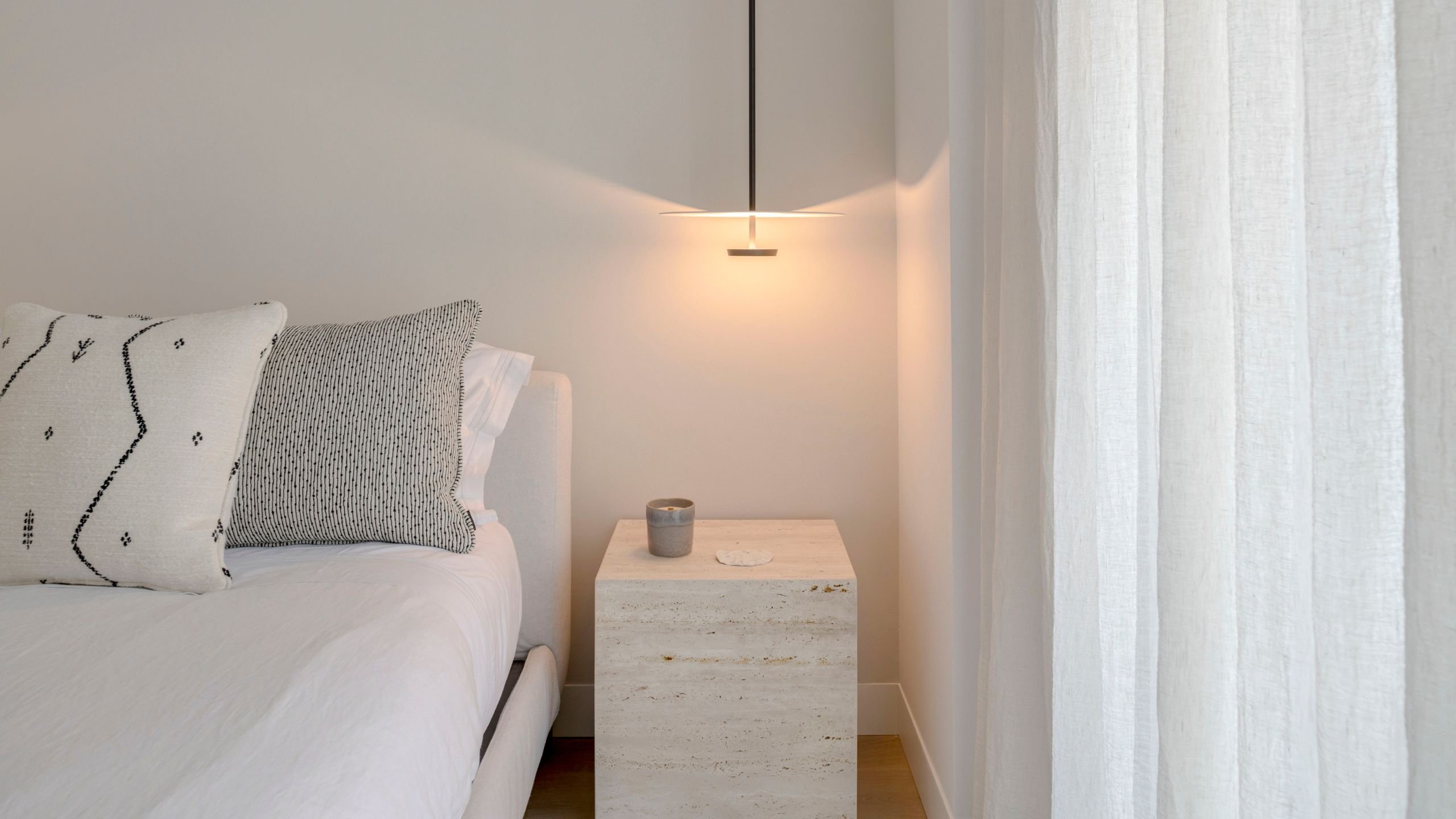 Mesinha de cabeceira em Travertino Romano com candeeiro suspenso minimalista ao lado da cama.
