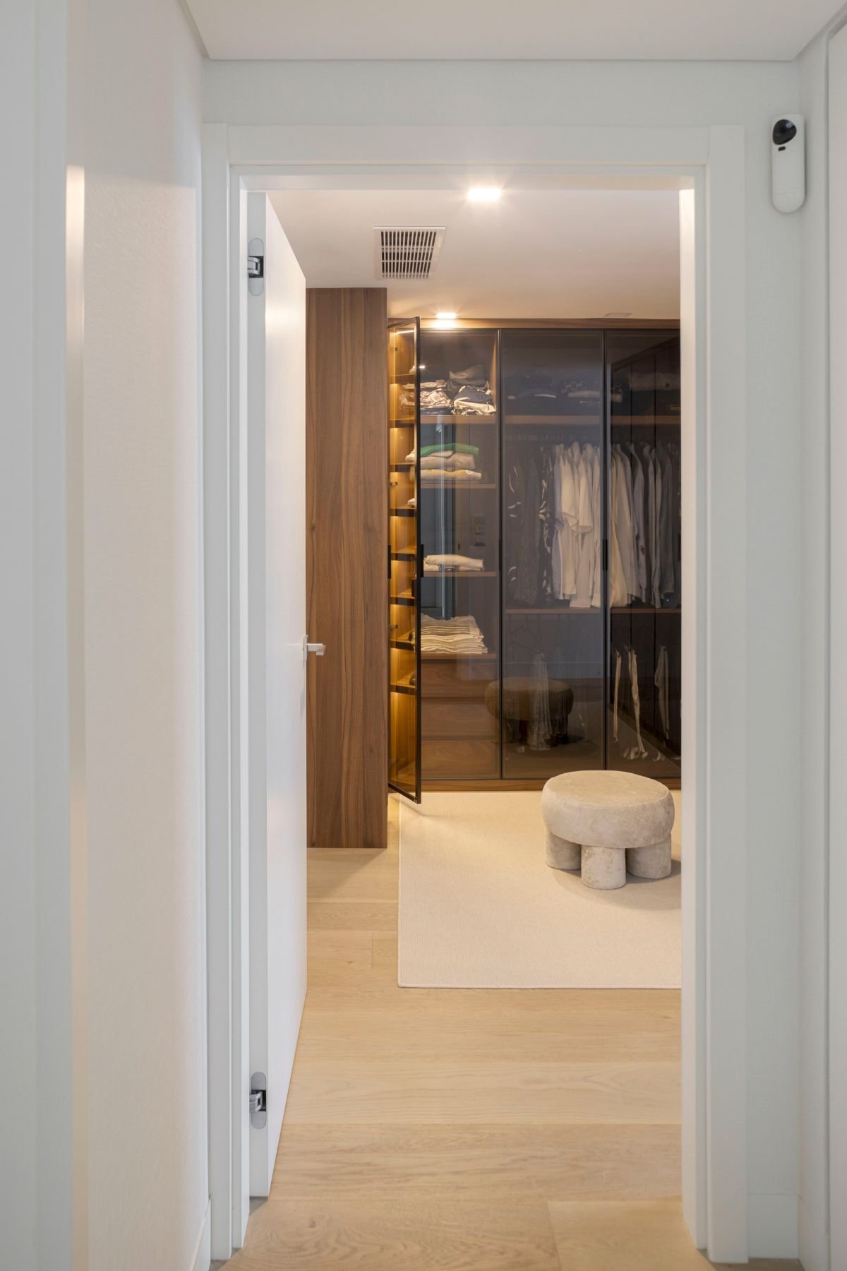 Vista do corredor de acesso ao closet com portas de vidro bronzeado e iluminação embutida.