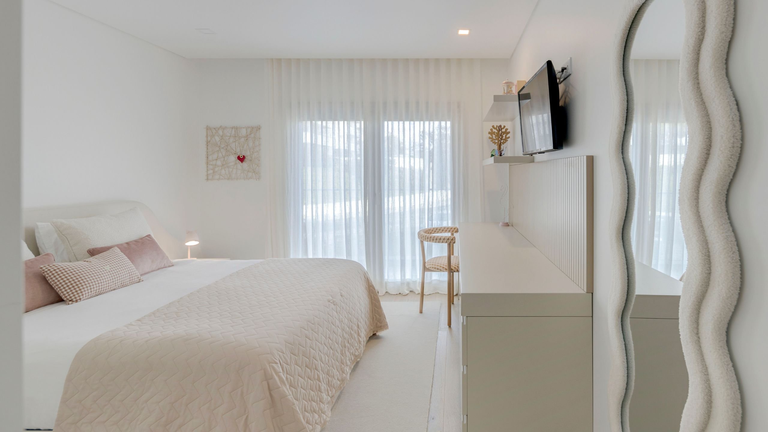 Quarto de criança em tons suaves com cama de casal, secretária integrada, espelho de moldura ondulada e ampla iluminação natural.