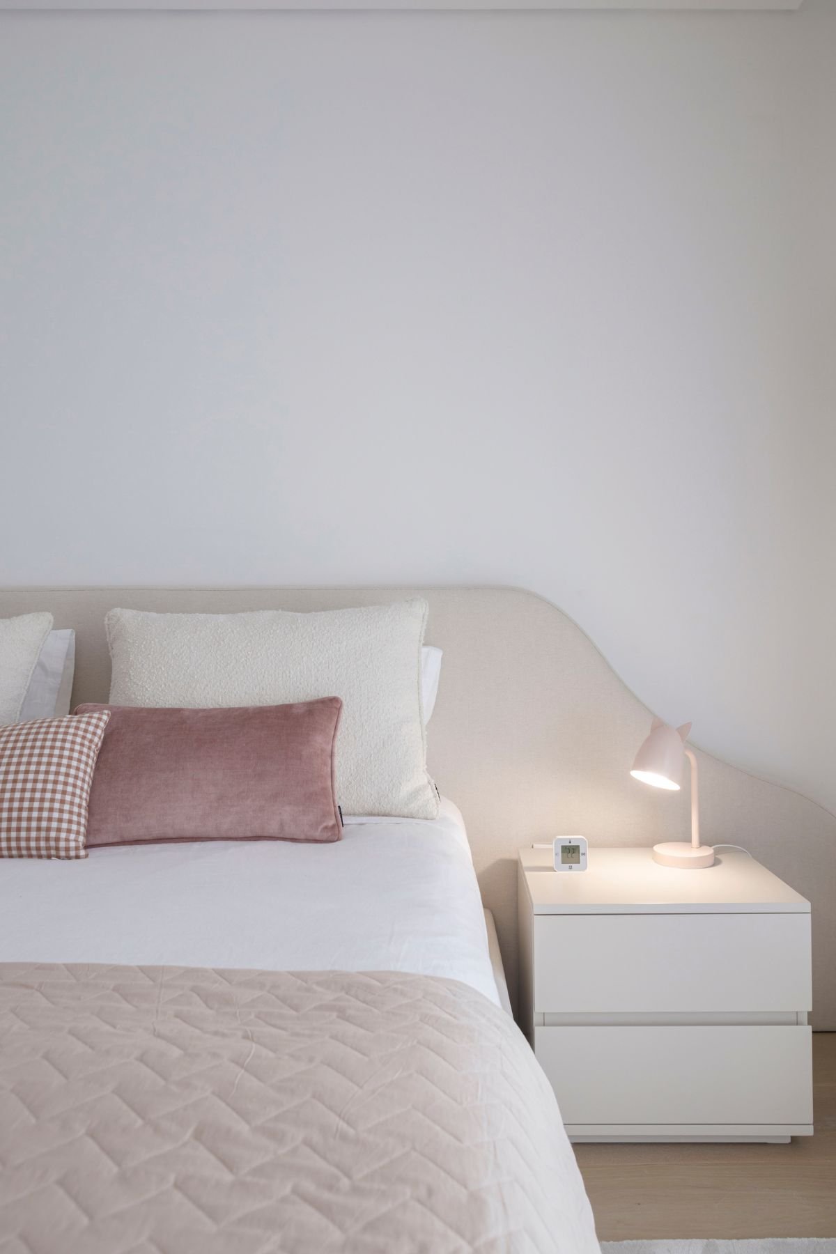Cabeceira de cama estofada com iluminação de apoio na mesa de cabeceira e almofadas em tons rosa e bege.