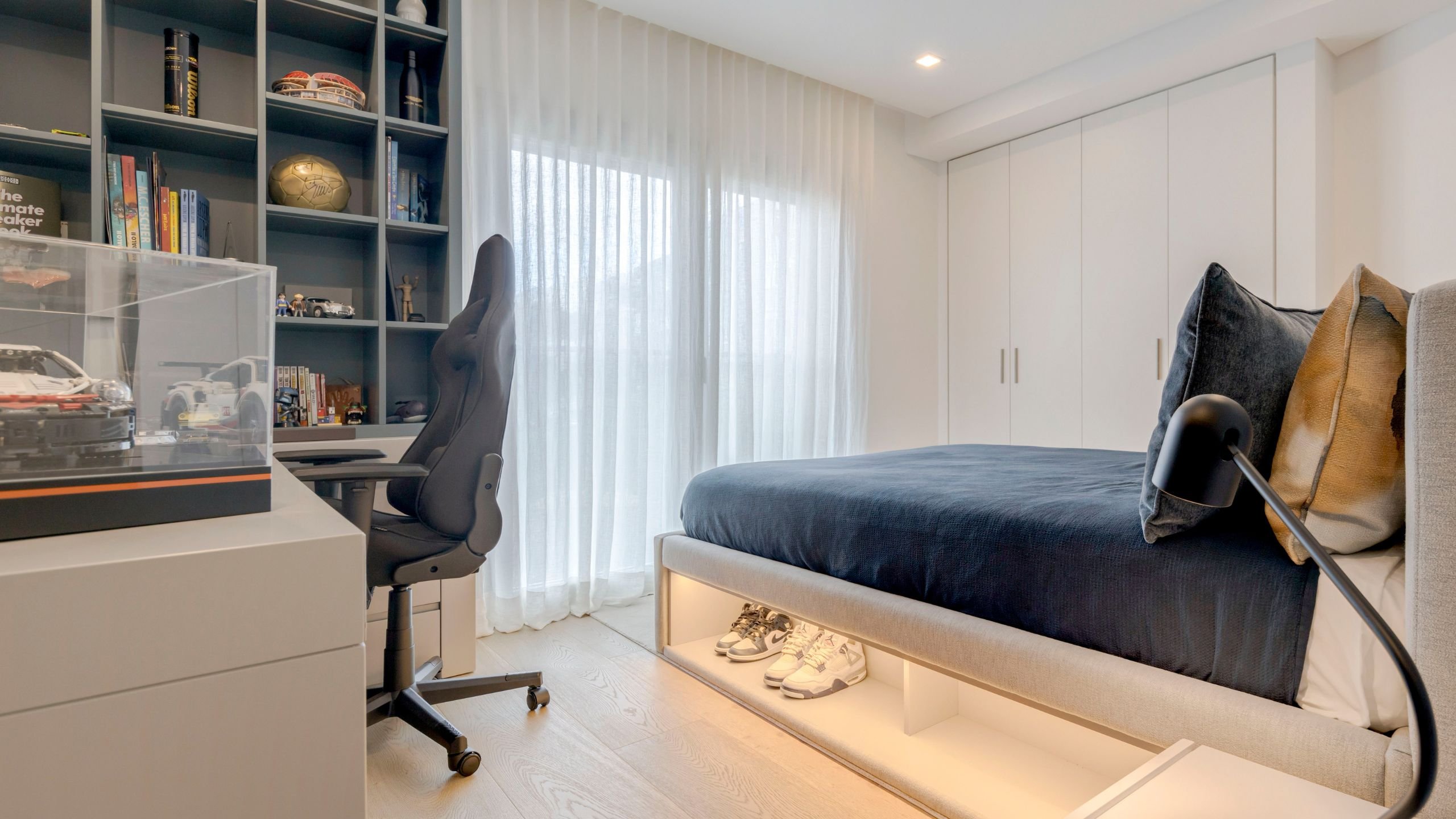 Quarto de adolescente em tons azuis com cama feita à medida com nicho iluminado para ténis, secretária e zona de estudo integrada.