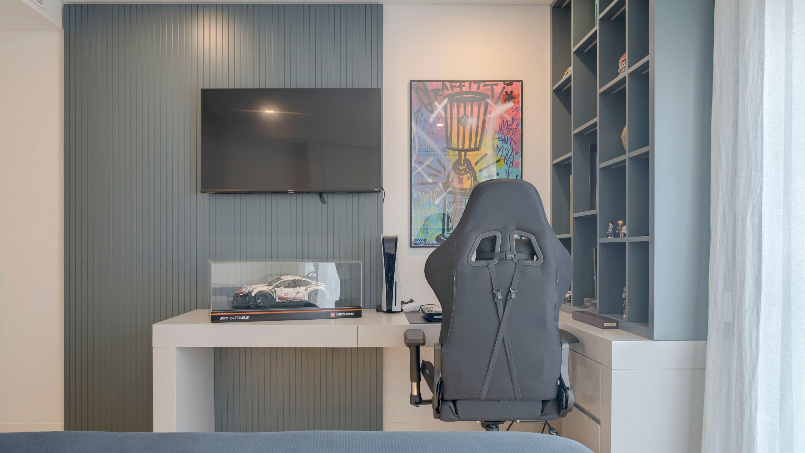 Vista frontal da bancada de trabalho com estante de livros, quadro urbano e suporte para consola de jogos.
