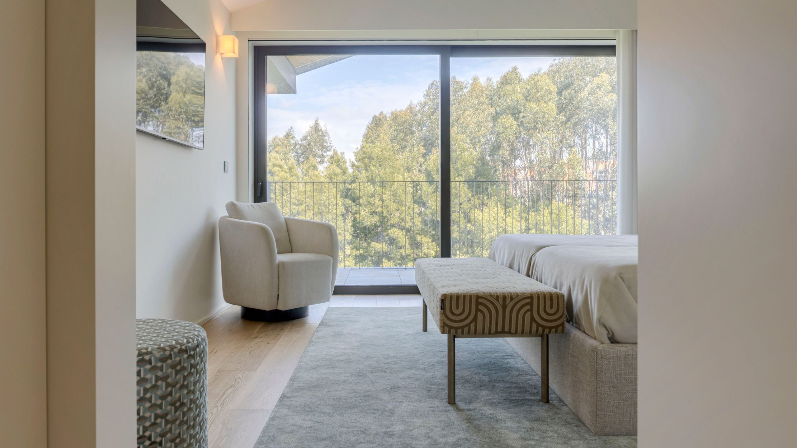 Vista panorâmica da suíte com banqueta aos pés da cama, cadeirão rotativo e pufe integrados num ambiente luminoso com vista para a natureza.