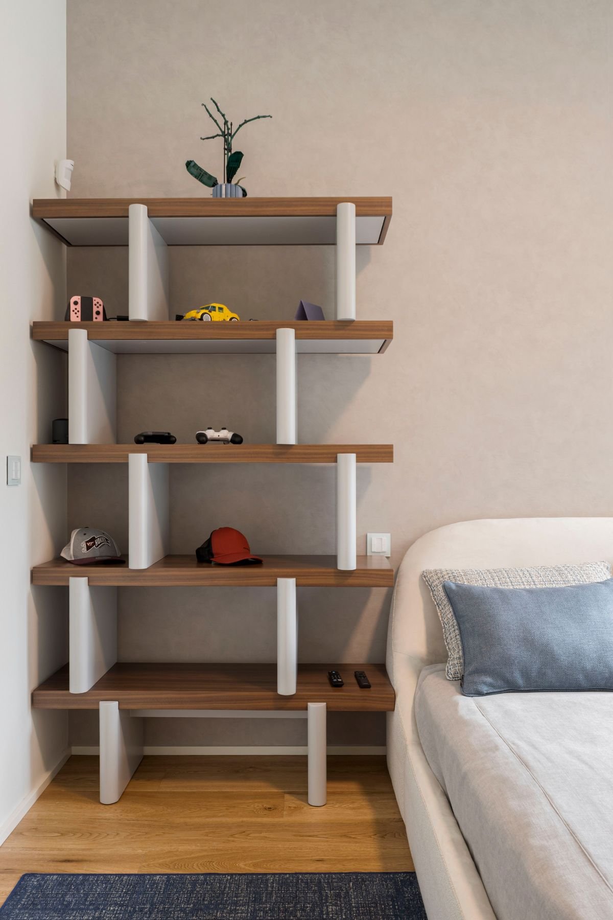 Estante para quarto em madeira Natur com prateleiras abertas para objetos pessoais, ao lado da cama de casal com cabeceira estofada.