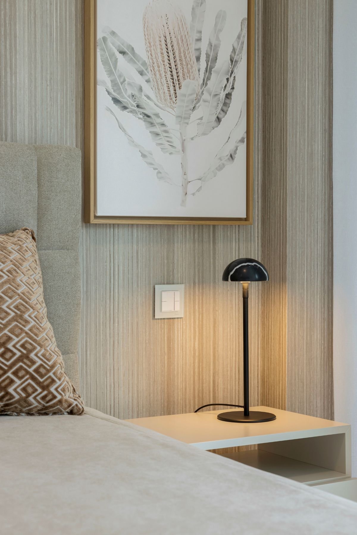 Pormenor da mesa de cabeceira branca com candeeiro de mesa minimalista e quadro decorativo sobre papel de parede texturizado.
