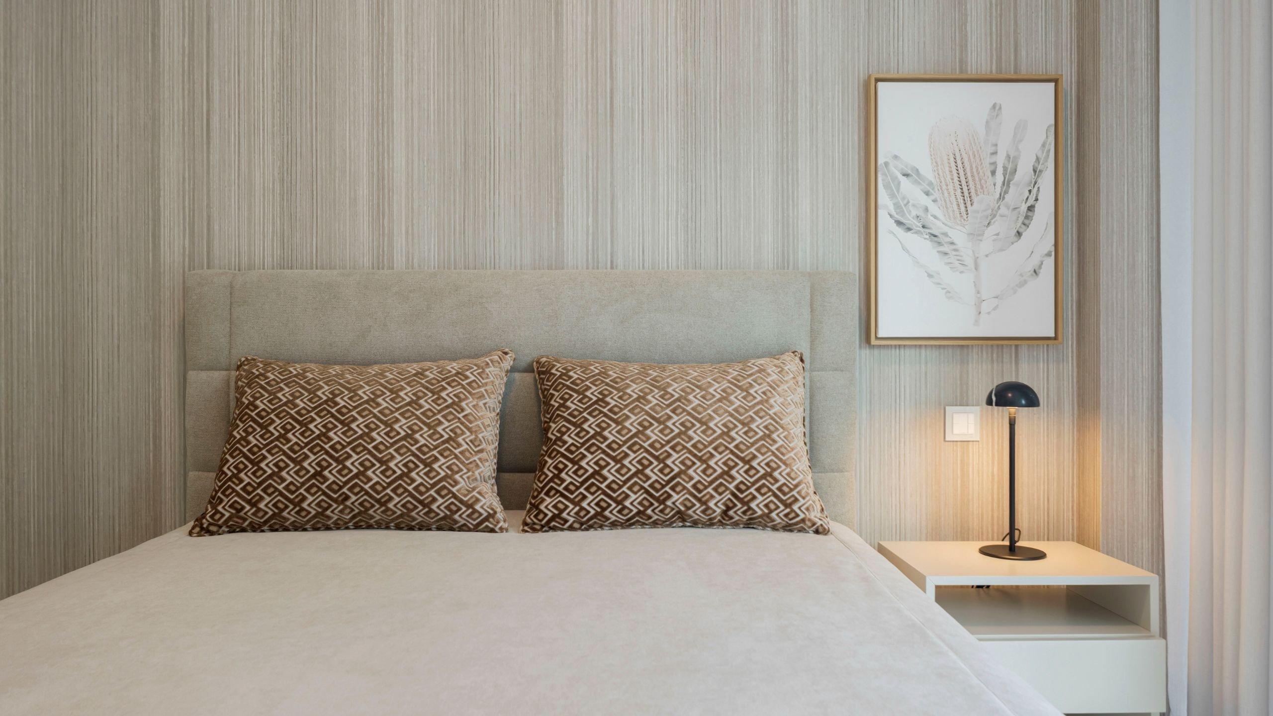 Cama de casal com cabeceira estofada em tom neutro, acompanhada por mesa de cabeceira moderna, candeeiro de mesa e quadro decorativo.